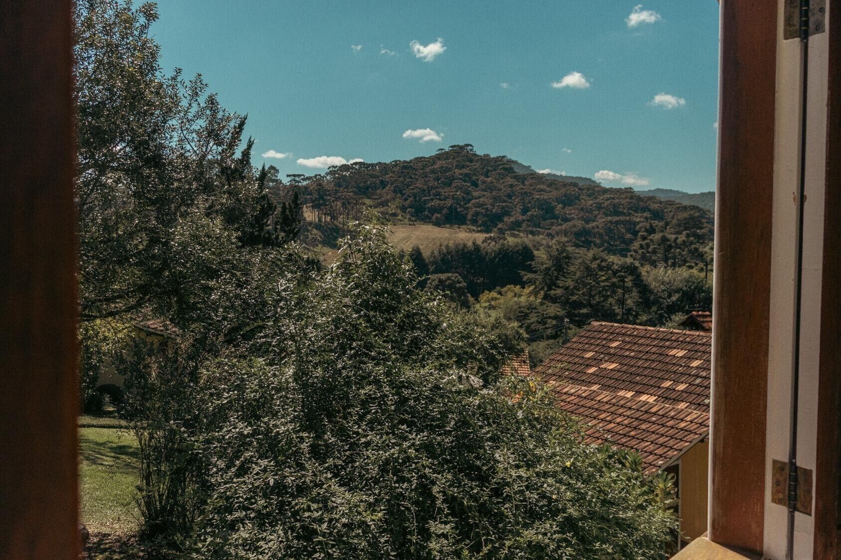 Une vue depuis une fenêtre donnant sur une colline, avec des arbres et un bâtiment doté d’un toit en tuiles.