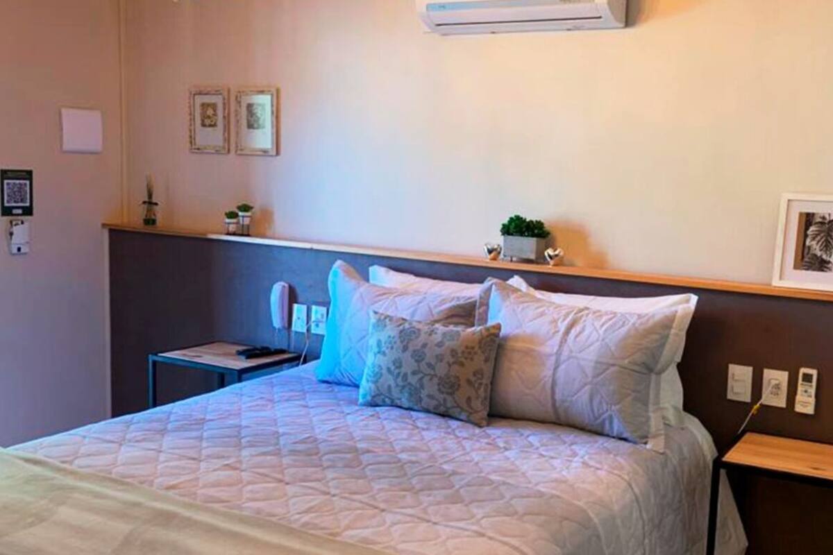 A neatly arranged hotel room with a bed, bedside tables, a wall-mounted air conditioner, and a small shelf with decorative items.