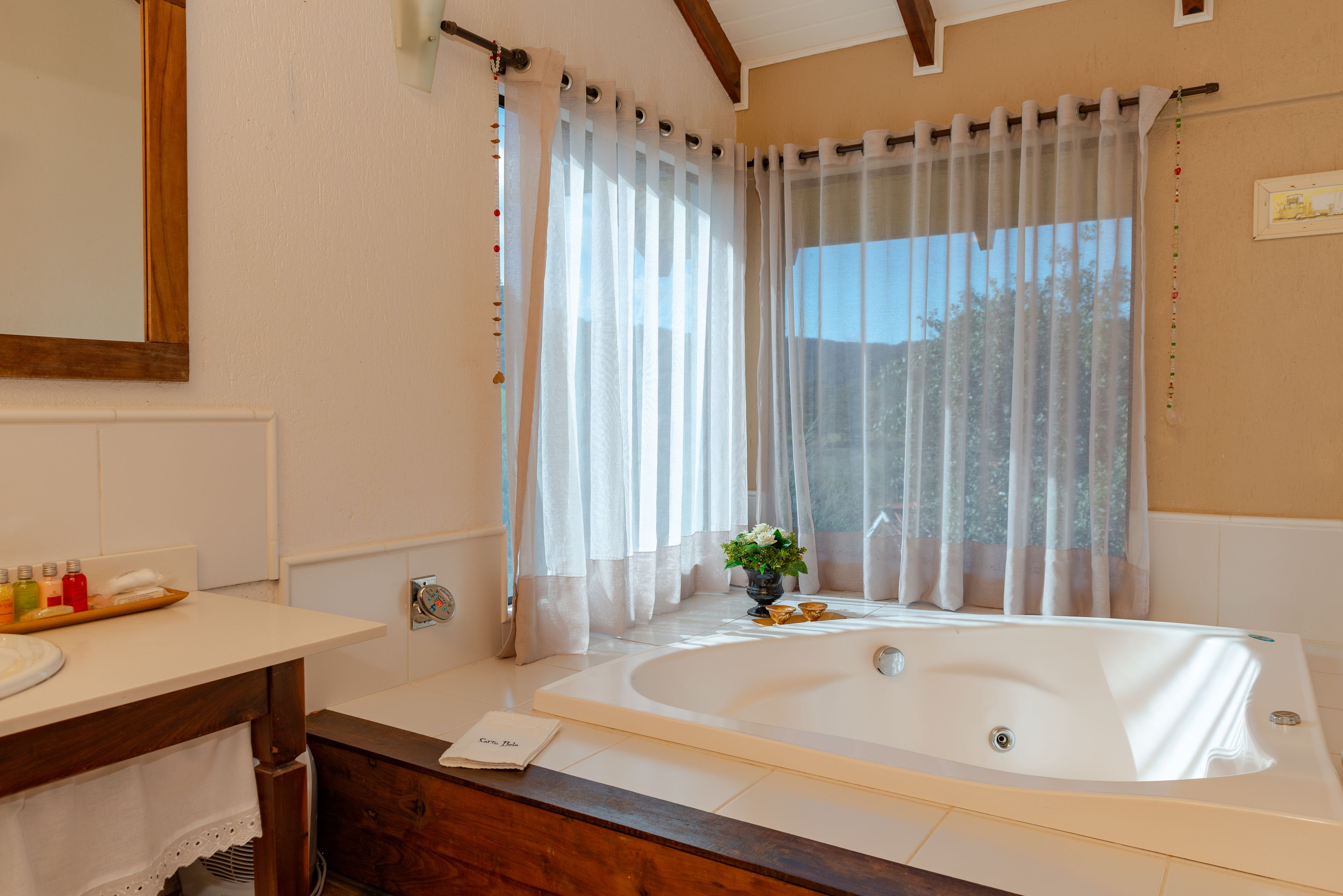 A modern bathroom with a large bathtub, a wooden vanity, a mirror, and a window with sheer curtains.