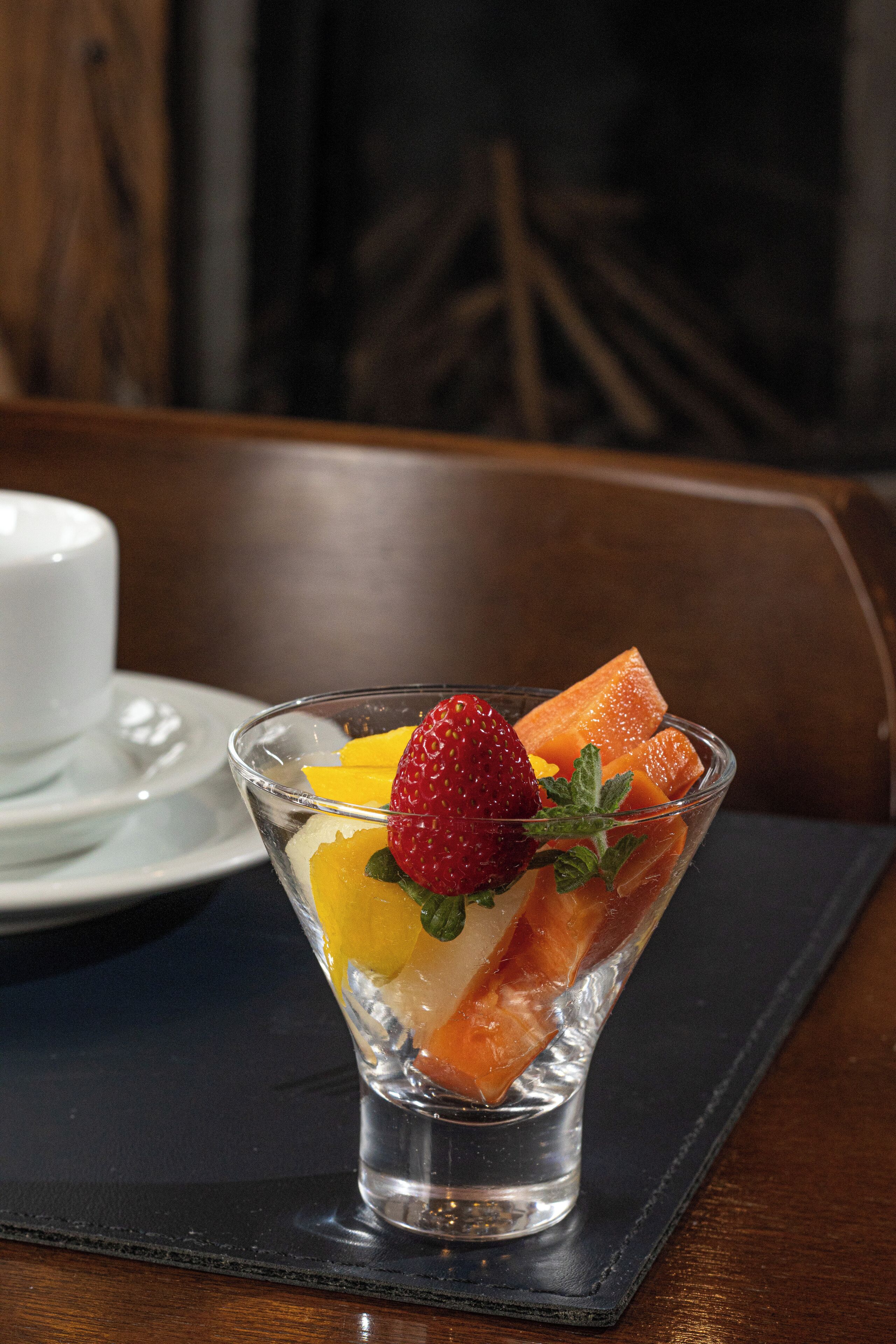 A clear glass dessert bowl on a dark placemat, containing fruit pieces and a strawberry, with a white cup and saucer in the background.