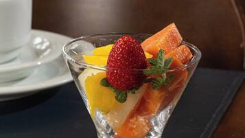 A clear glass dessert bowl on a dark placemat, containing fruit pieces and a strawberry, with a white cup and saucer in the background.