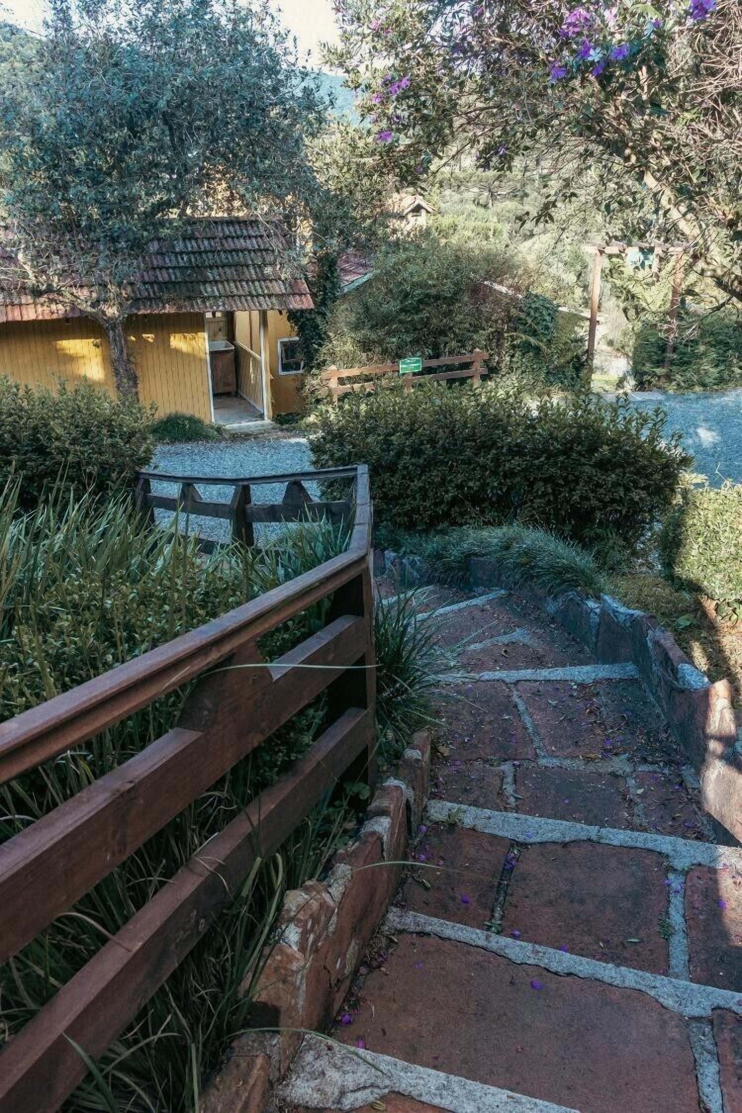 A stone pathway leading to a wooden structure with a tiled roof, surrounded by lush greenery and a wooden fence.