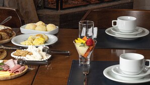 Une table à manger est dressée avec des assiettes remplies de nourriture, un verre d’eau et une tasse. Au fond, on aperçoit une cheminée où brûle un feu.