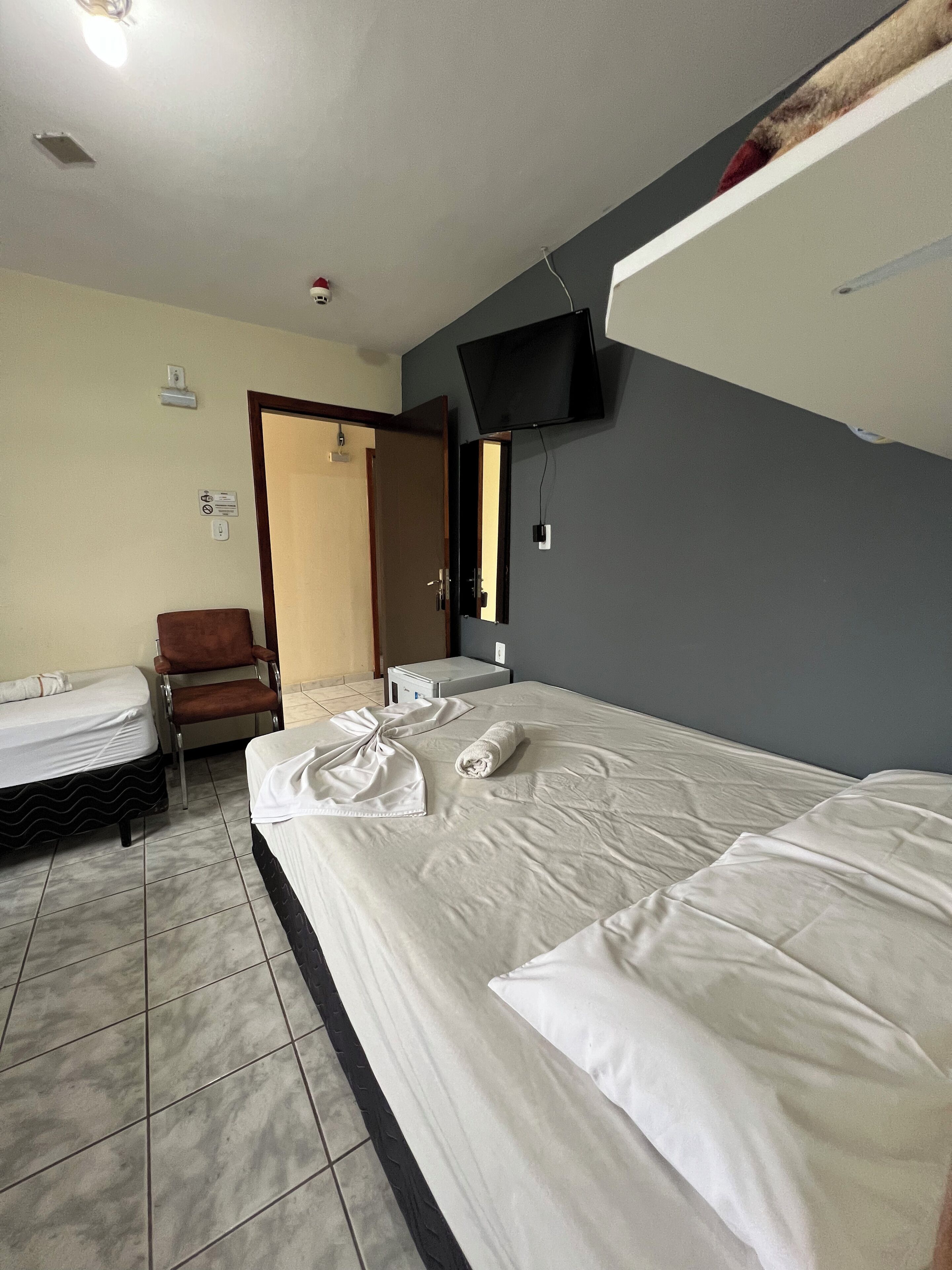 Quarto de hotel com cama, uma cadeira, escrivaninha, TV de tela plana na parede e banheiro com pia e espelho.