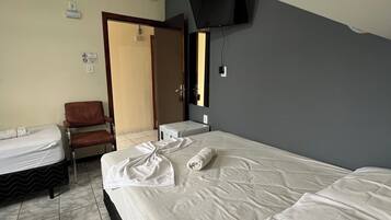 Quarto de hotel com cama, uma cadeira, escrivaninha, TV de tela plana na parede e banheiro com pia e espelho.