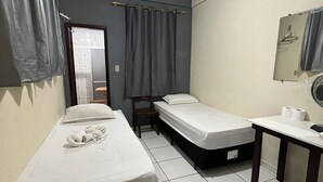 A small, clean room with two single beds, a small table, a chair, and a window with curtains.