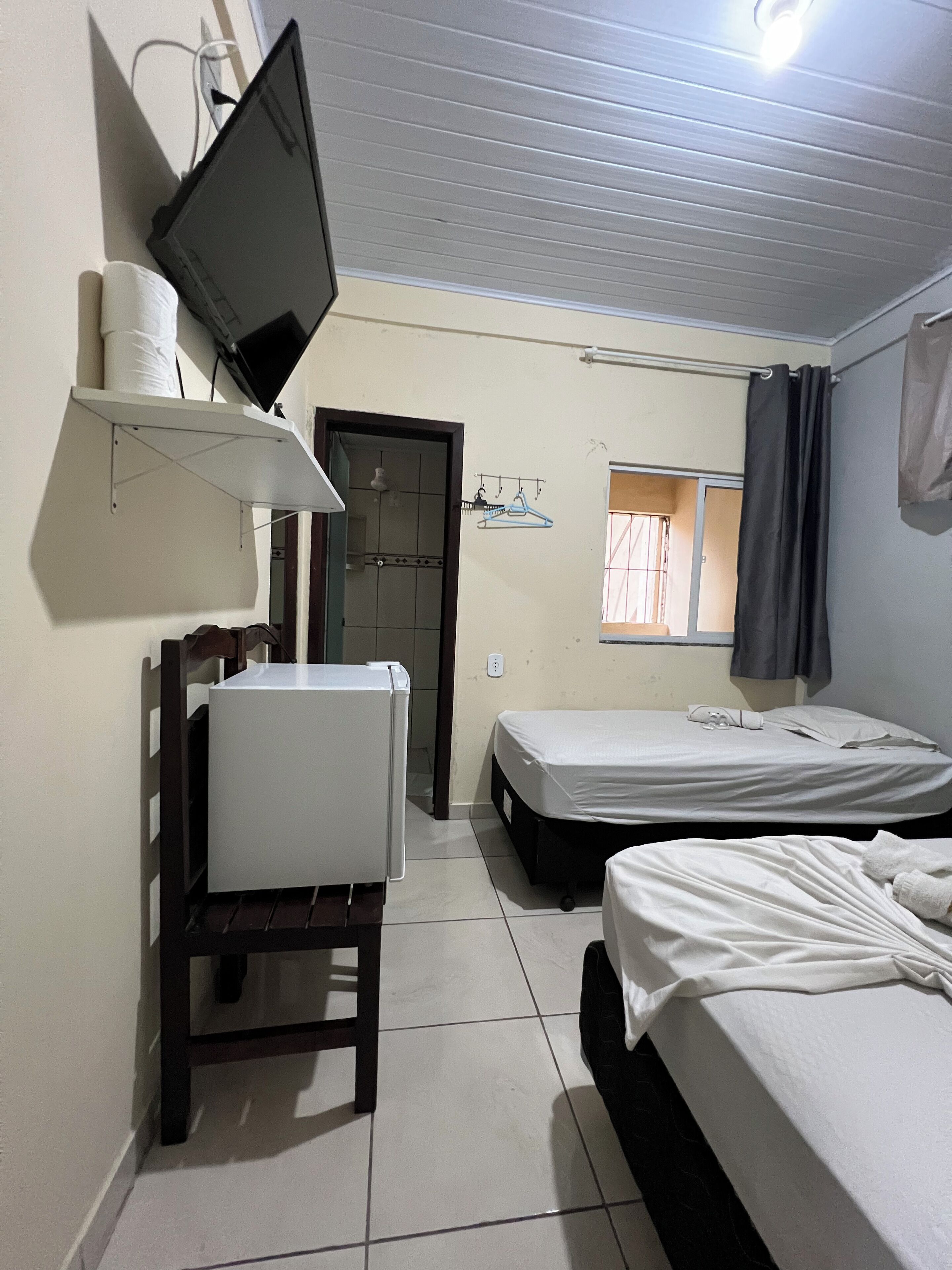A room with two beds, a TV mounted on the wall, a small window with curtains, and a door leading to another room.