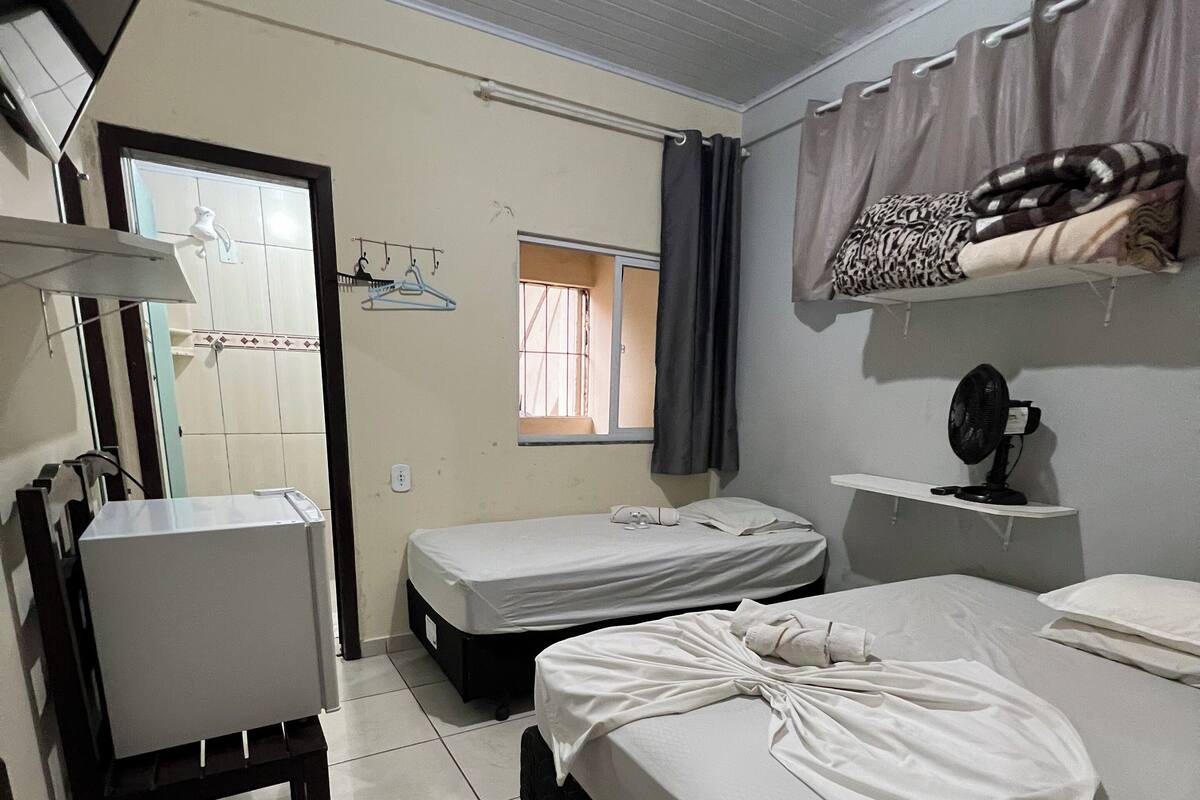 A hotel room with a bed, a small table, a chair, a window, a fan, and a towel on the bed.