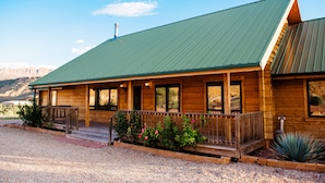 A wooden cabin with a green metal roof, a porch with a railing, and a gravel driveway.