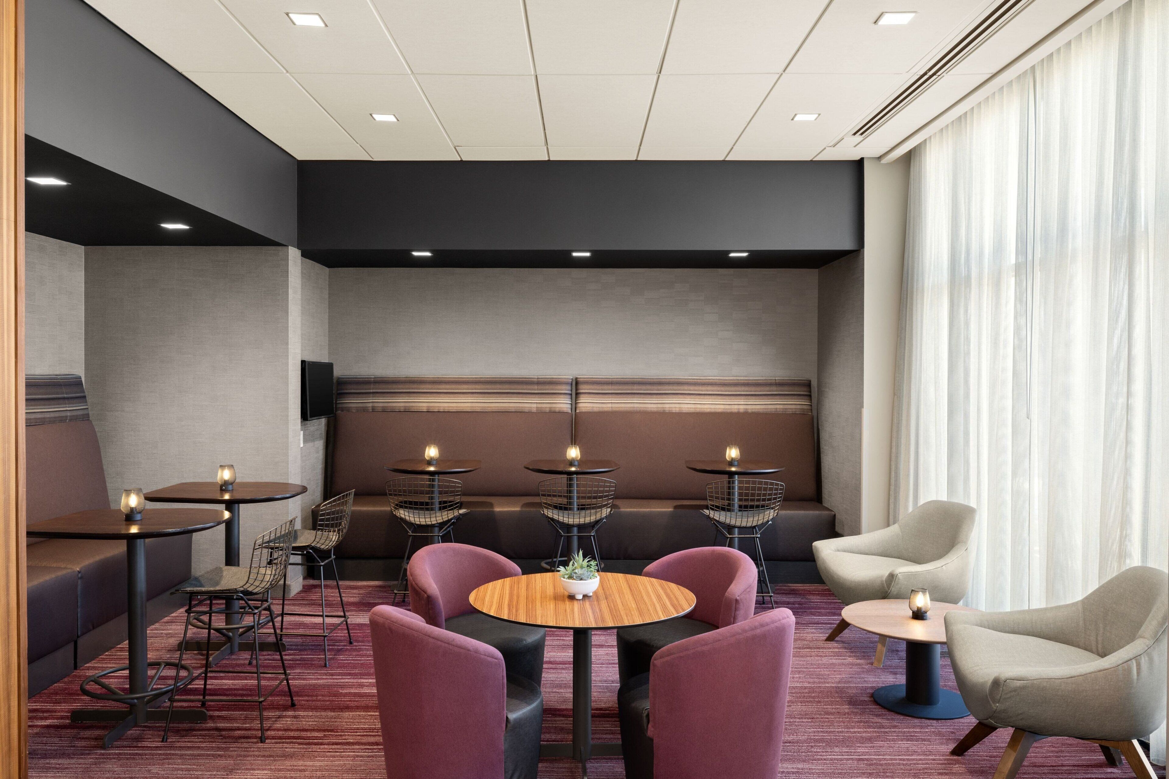A modern hotel room with a seating area featuring round tables, cushioned chairs, and a large window with curtains.