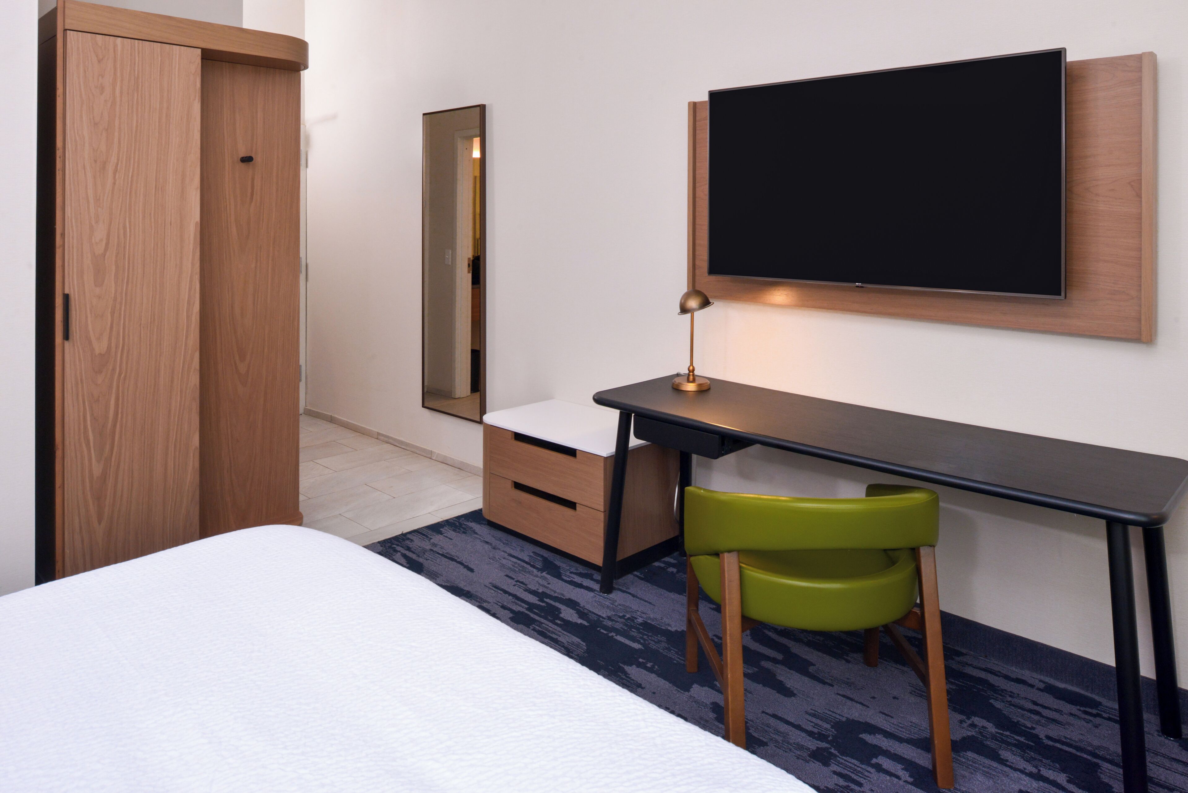 A hotel room with a bed, a desk with a chair, a TV, a wardrobe, and a mirror.