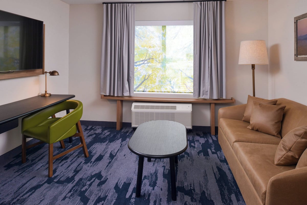Une chambre d’hôtel équipée d’un bureau, d’un fauteuil vert, d’un canapé et d’une fenêtre avec des rideaux.