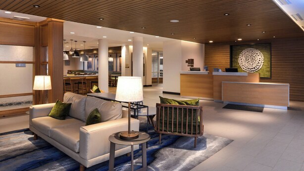 Un moderno vestíbulo de hotel con una recepción, una zona de estar con sofá y sillones, y un comedor al fondo.