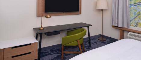 Une chambre d’hôtel comprenant un lit, un bureau avec une chaise, un téléviseur fixé au mur, une lampe et une fenêtre avec des rideaux.