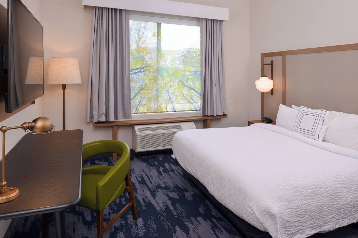 Une chambre d’hôtel comprenant un lit, un bureau avec une lampe, une chaise, une fenêtre avec des rideaux et une table de chevet avec une lampe.