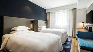 A hotel room with two beds, a desk with a chair, a flat-screen TV, and a window with curtains.