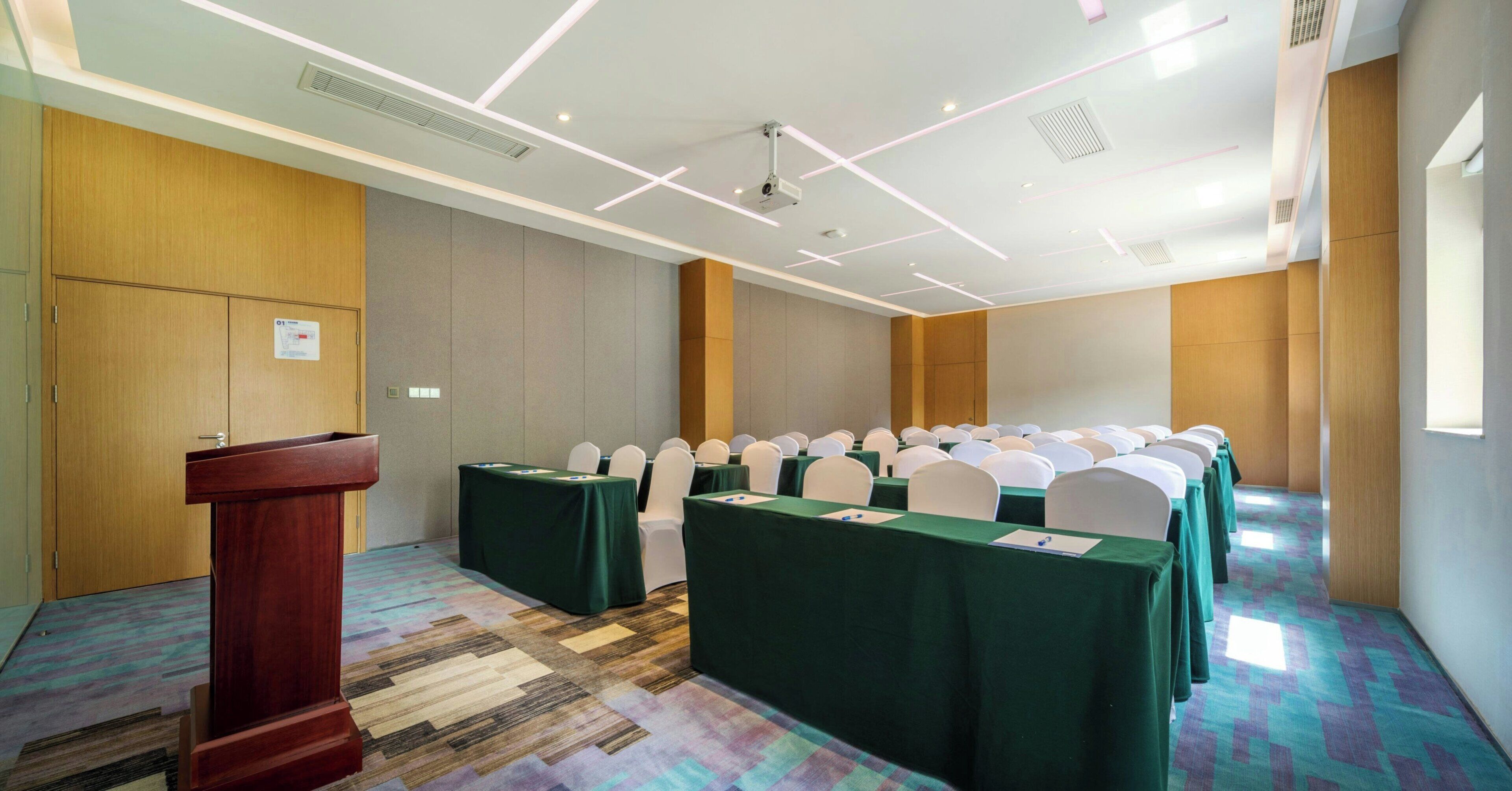 A conference room with a podium, rows of tables with green tablecloths, white chairs, and a patterned carpet.