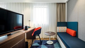 A modern hotel room with a flat-screen TV, a desk with a chair, a small round table, and a sofa with cushions.