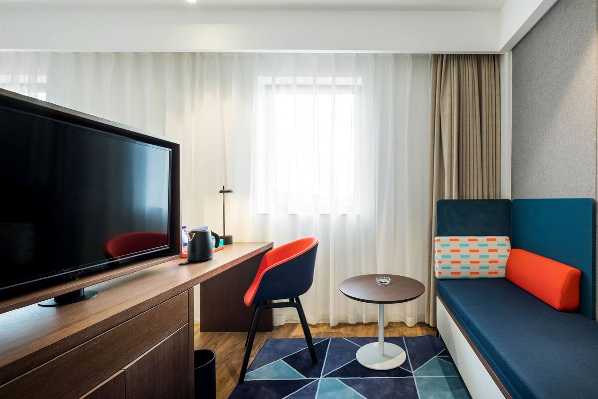 Una camera d'albergo moderna con televisione a schermo piatto, una scrivania con sedia, un piccolo tavolino rotondo e un divano con cuscini.