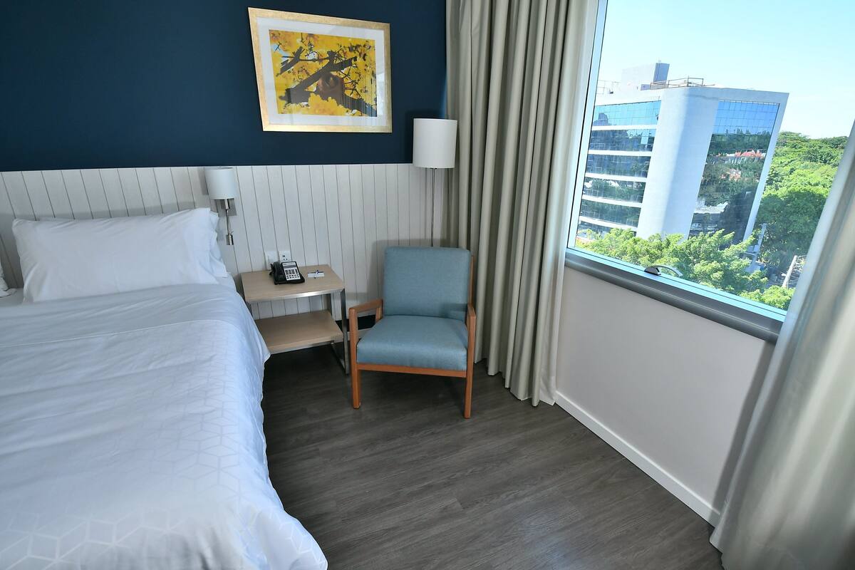 A hotel room with a bed, a desk with a lamp, a chair, and a window with a view of buildings and trees.