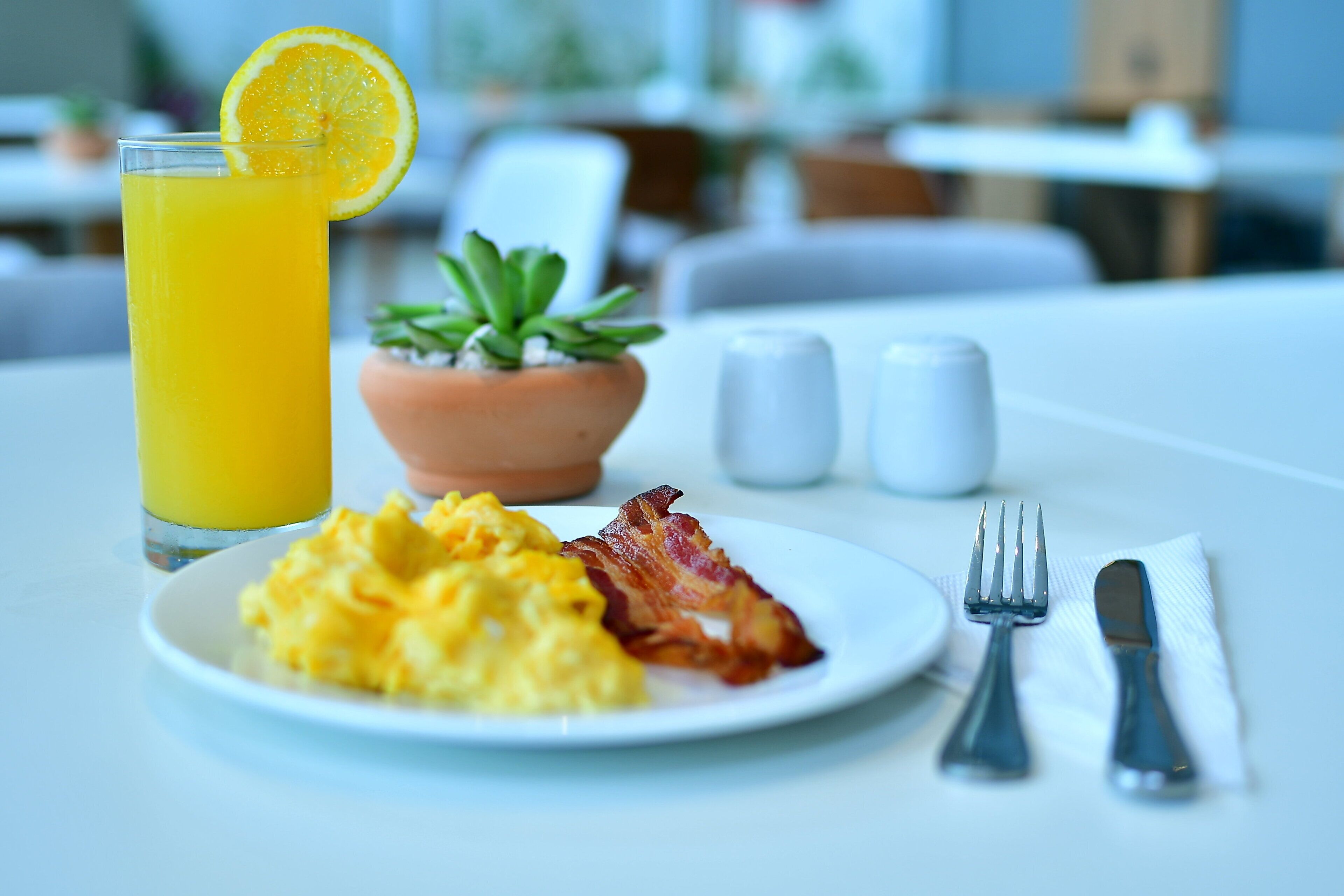 A breakfast setting with scrambled eggs, bacon, a glass of orange juice, a small potted plant, salt and pepper shakers, and cutlery on a table.