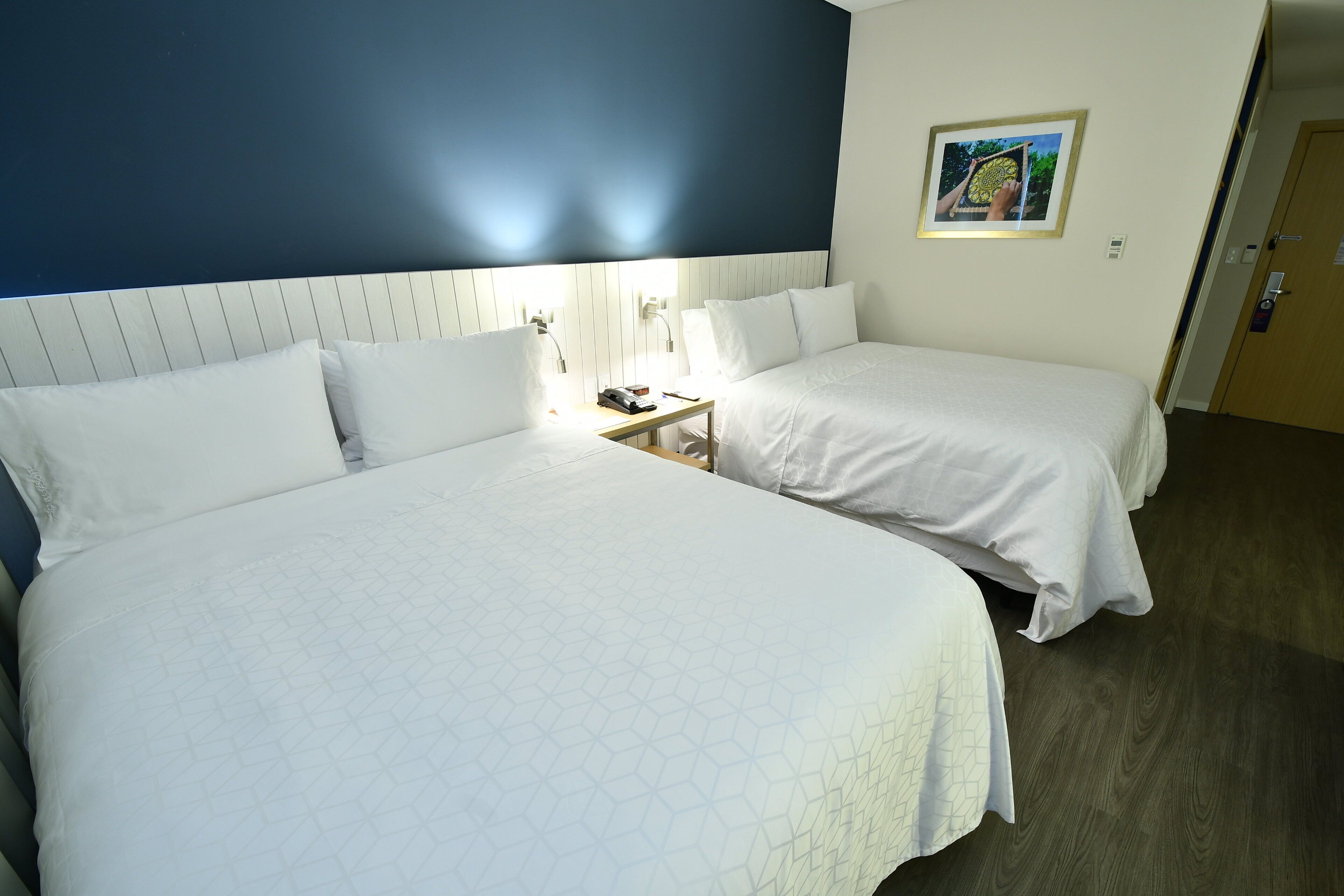 Hotellihuone, jossa on kaksi sänkyä, yöpöytä lampun kanssa ja seinällä oleva kehystetty kuva.