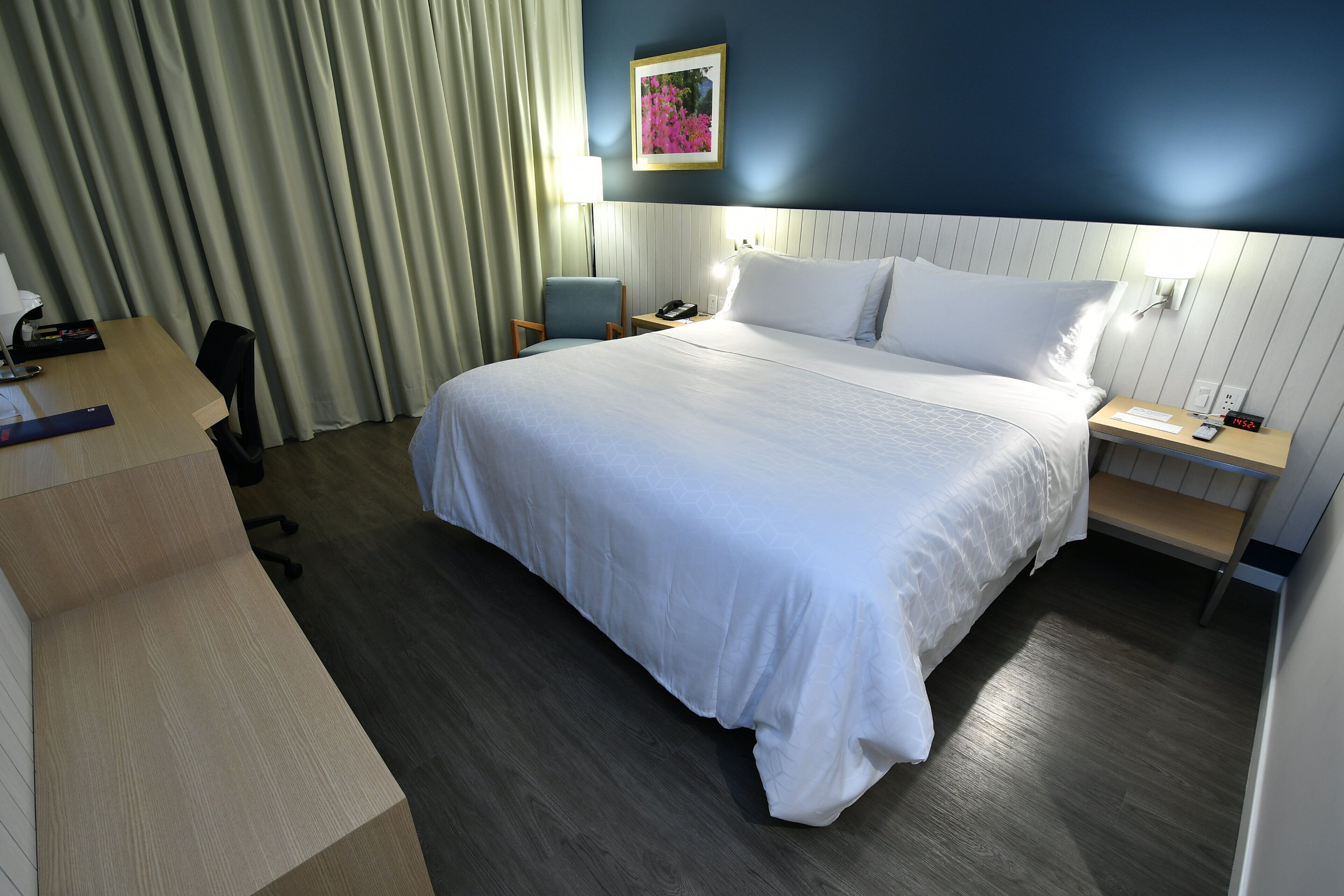 A hotel room with a large bed, a desk, a chair, a nightstand, a lamp, and a picture on the wall.