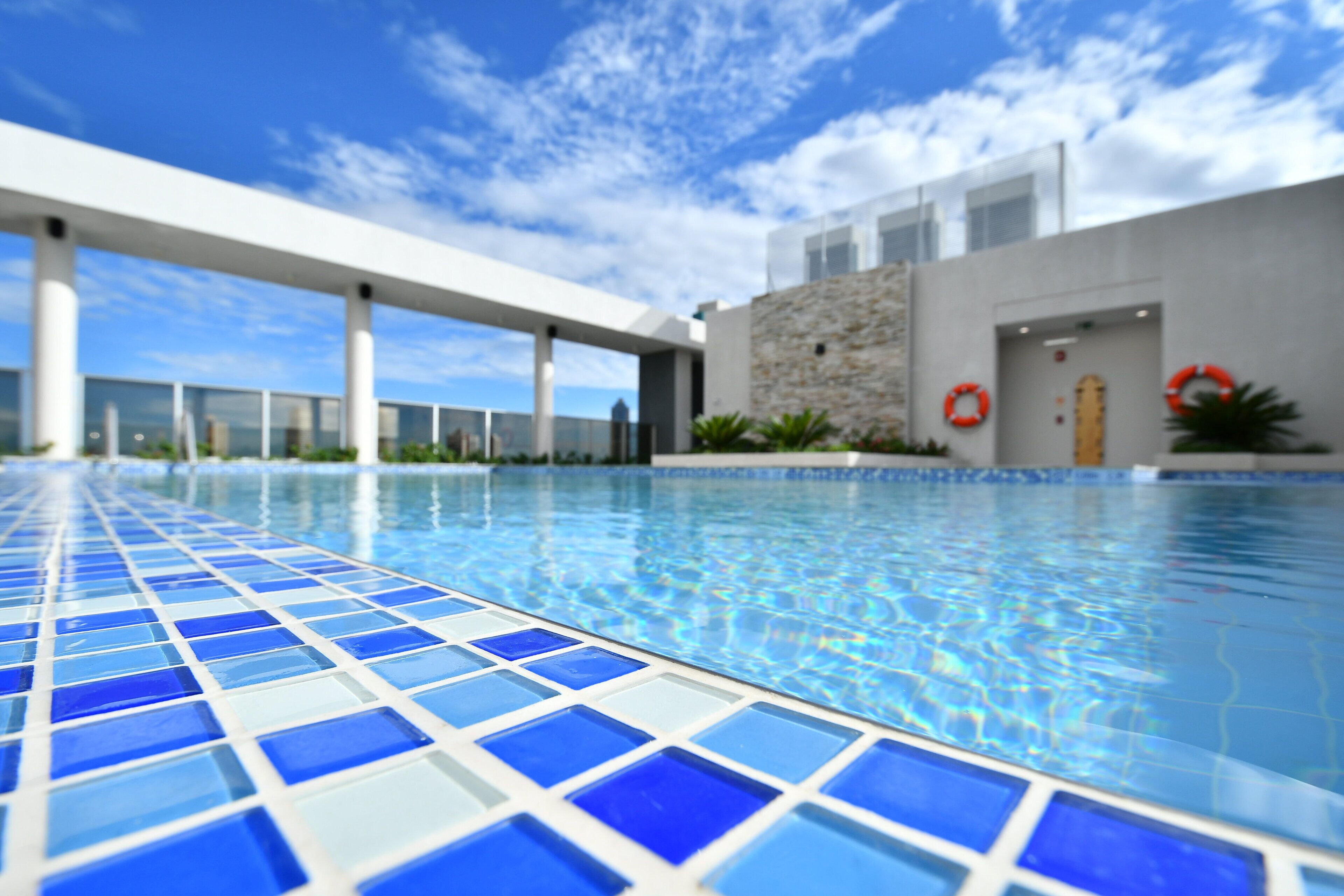 Uma piscina com azulejos azuis e brancos, um edifício moderno com fachada de pedra e um céu azul límpido.