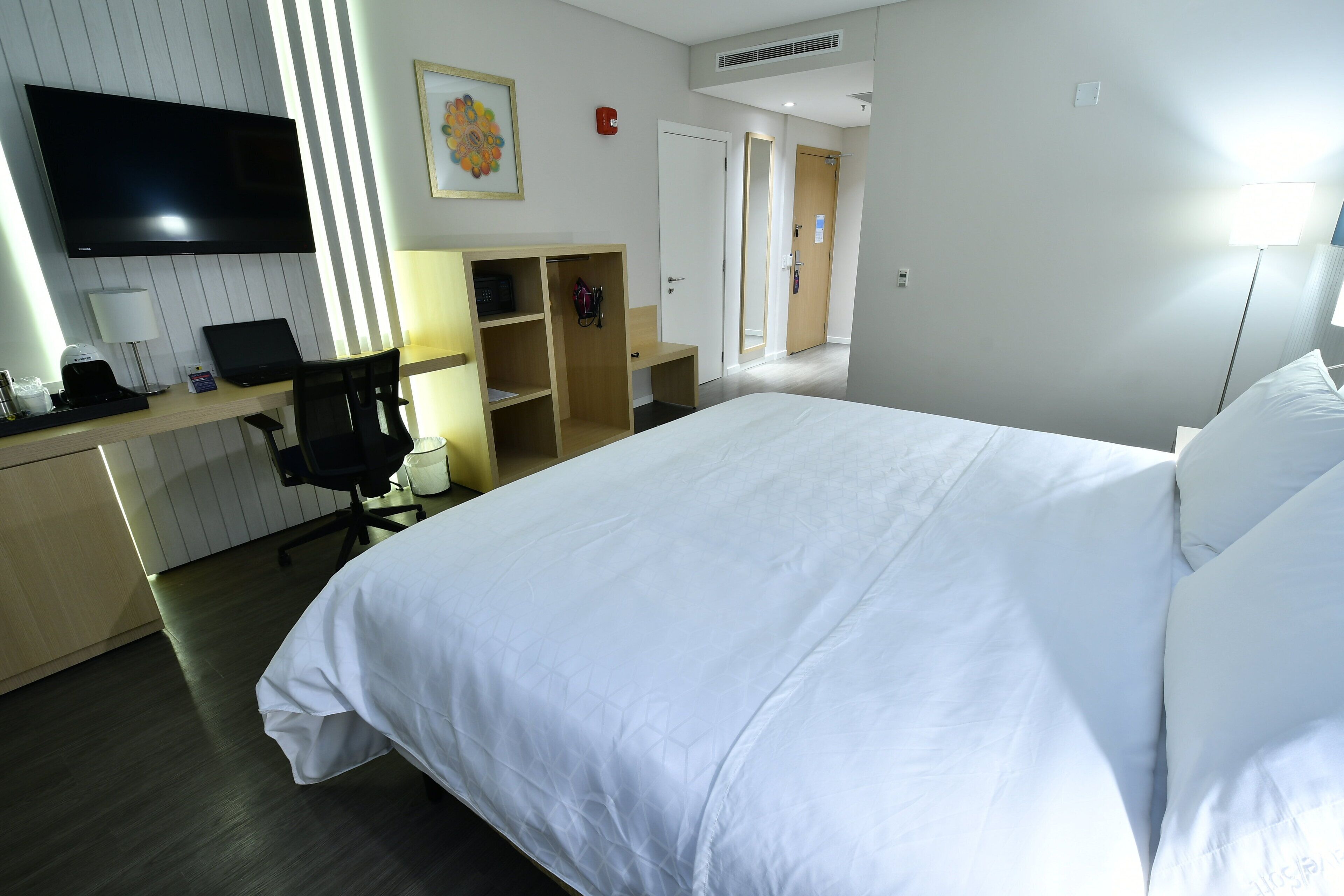 A hotel room with a large bed, a desk with a laptop, a chair, a TV, and a wardrobe.