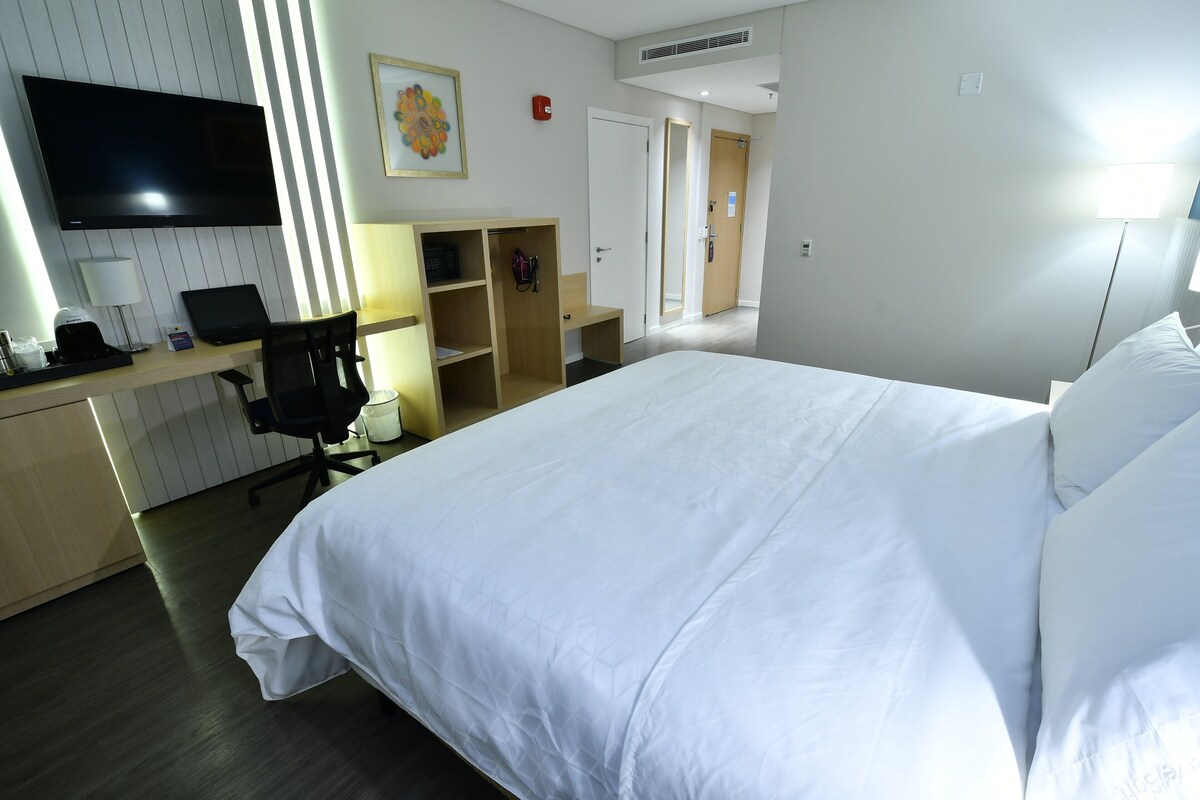 A hotel room with a large bed, a desk with a laptop, a chair, a TV, and a wardrobe.