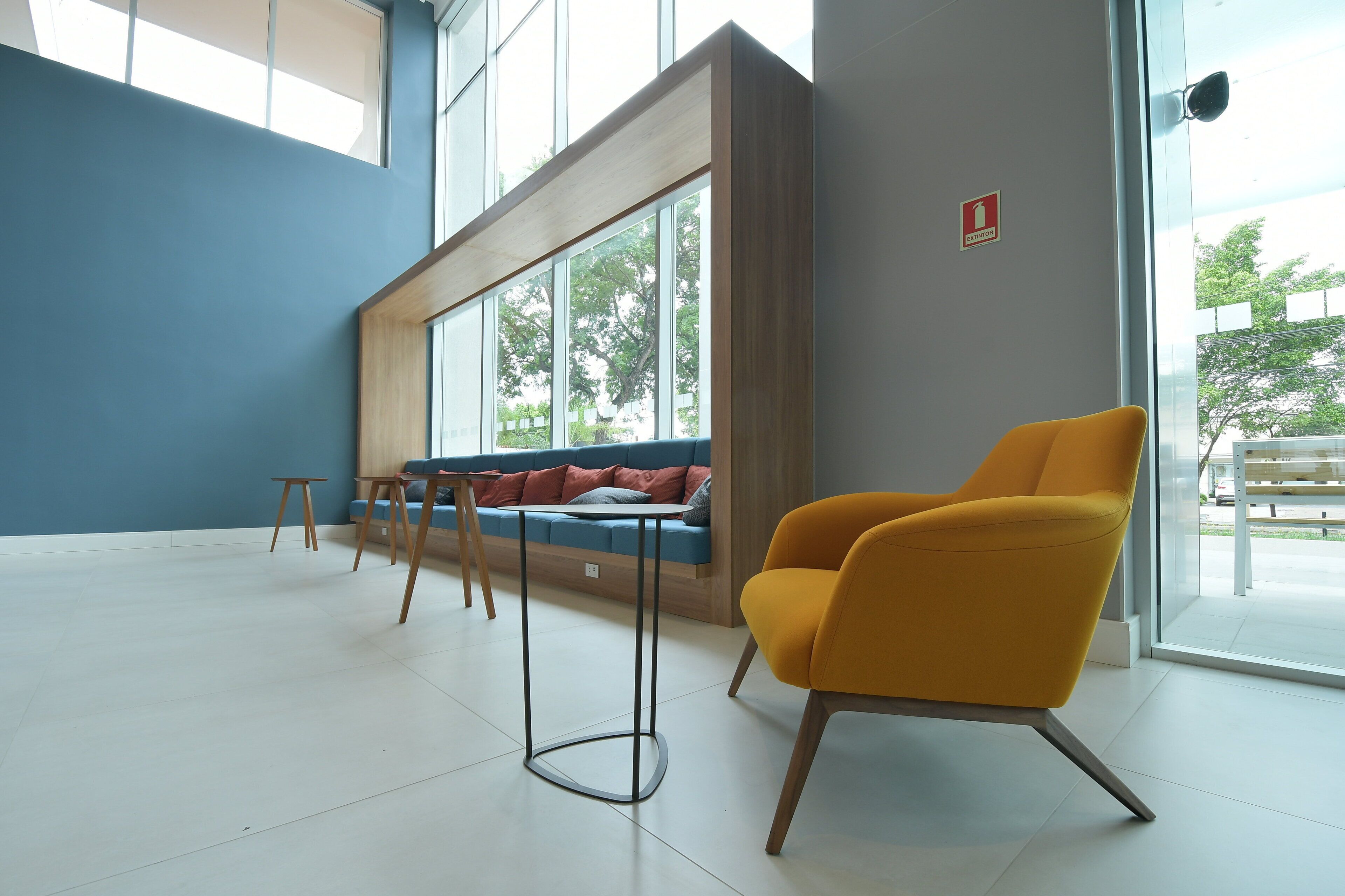 Ein modernes Interieur mit blauer Wand, einer Holzbank mit Kissen, einem gelben Sessel und einem Fenster mit Blick auf Bäume.