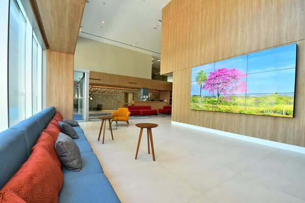A modern lobby with a blue sofa, red and grey cushions, wooden flooring, and a large wall-mounted TV displaying a pink tree.
