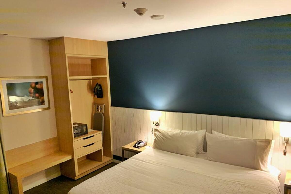 A hotel room with a large bed, a bedside table with a lamp, a wardrobe, and a small desk with a chair.