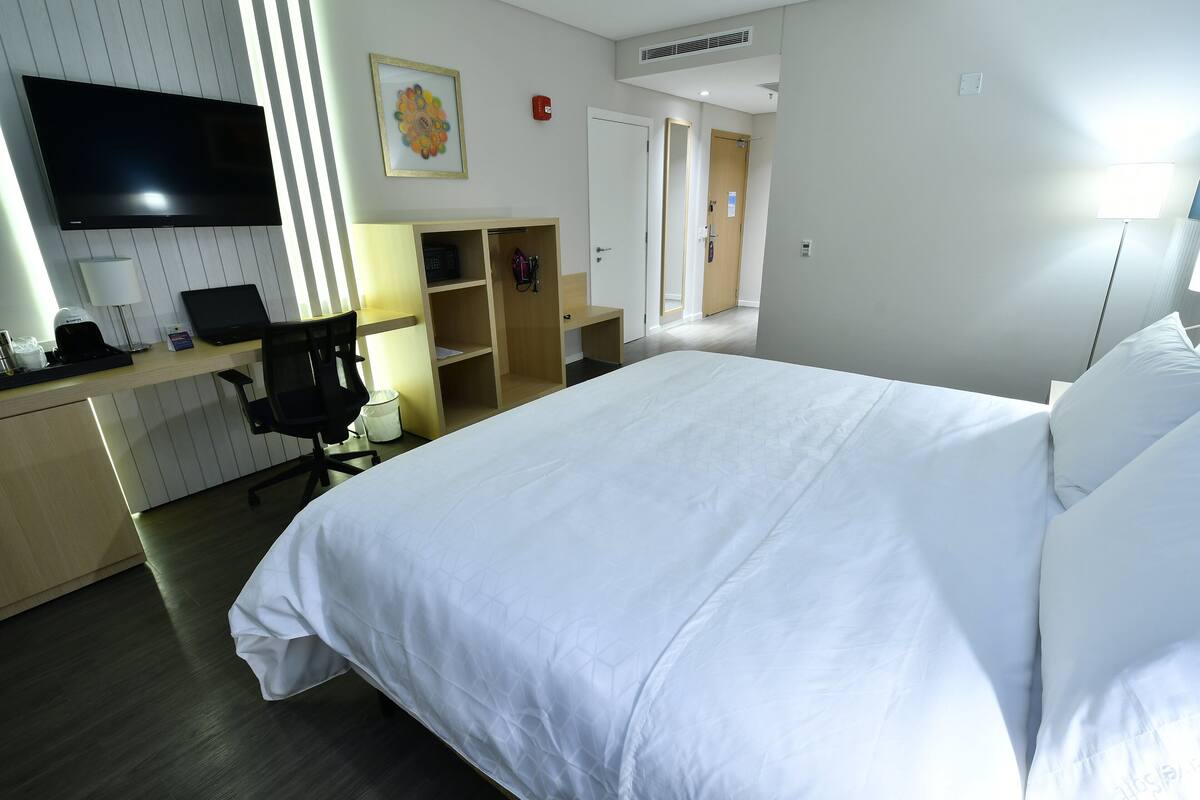 A hotel room with a large bed, a desk with a laptop, a chair, a TV, and a wardrobe.