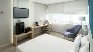 A hotel room with a bed, a desk with a laptop, a chair, a TV, and a window with blinds.