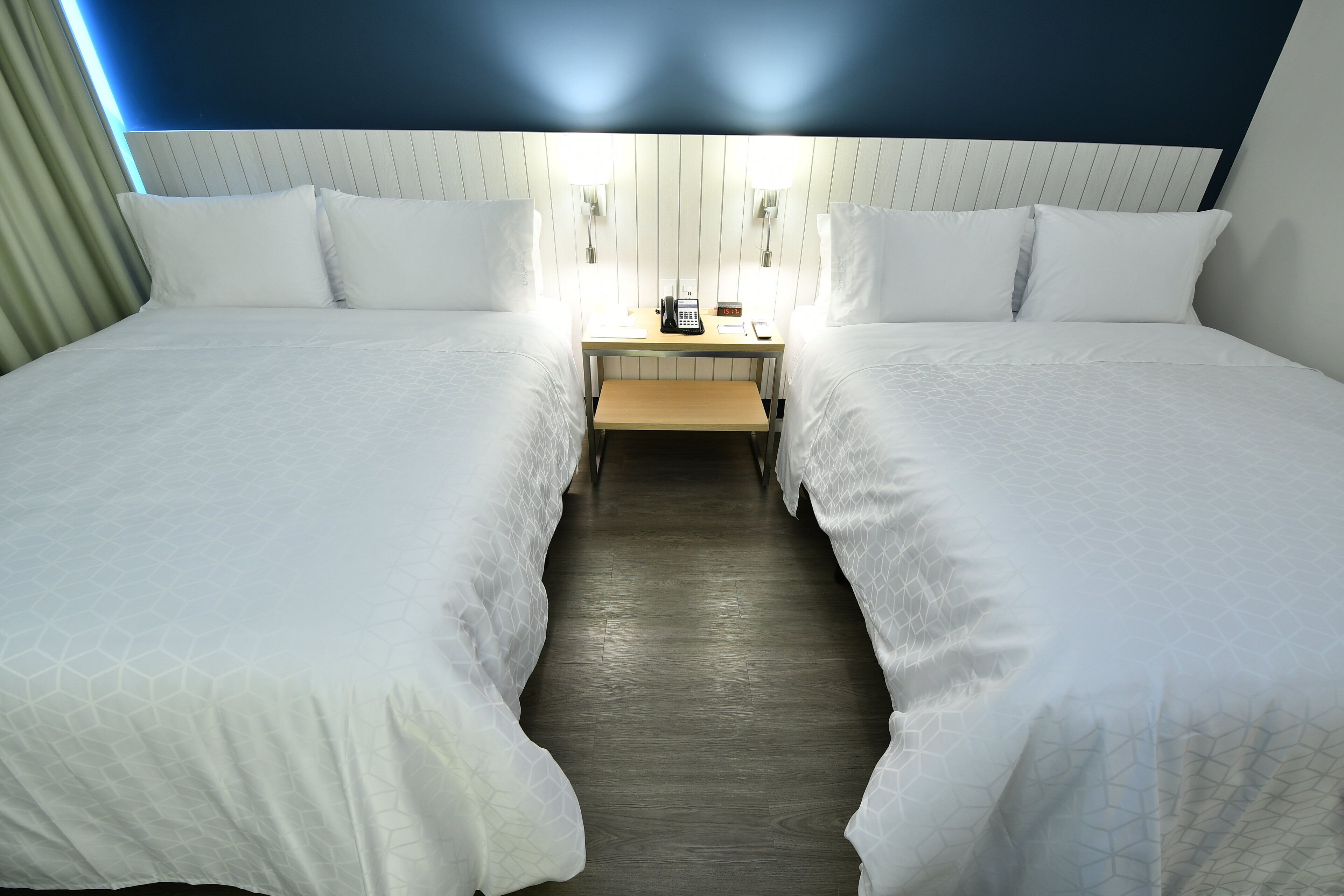 Habitación de hotel con dos camas, una mesita de noche con teléfono y lámpara, y un banco de madera.