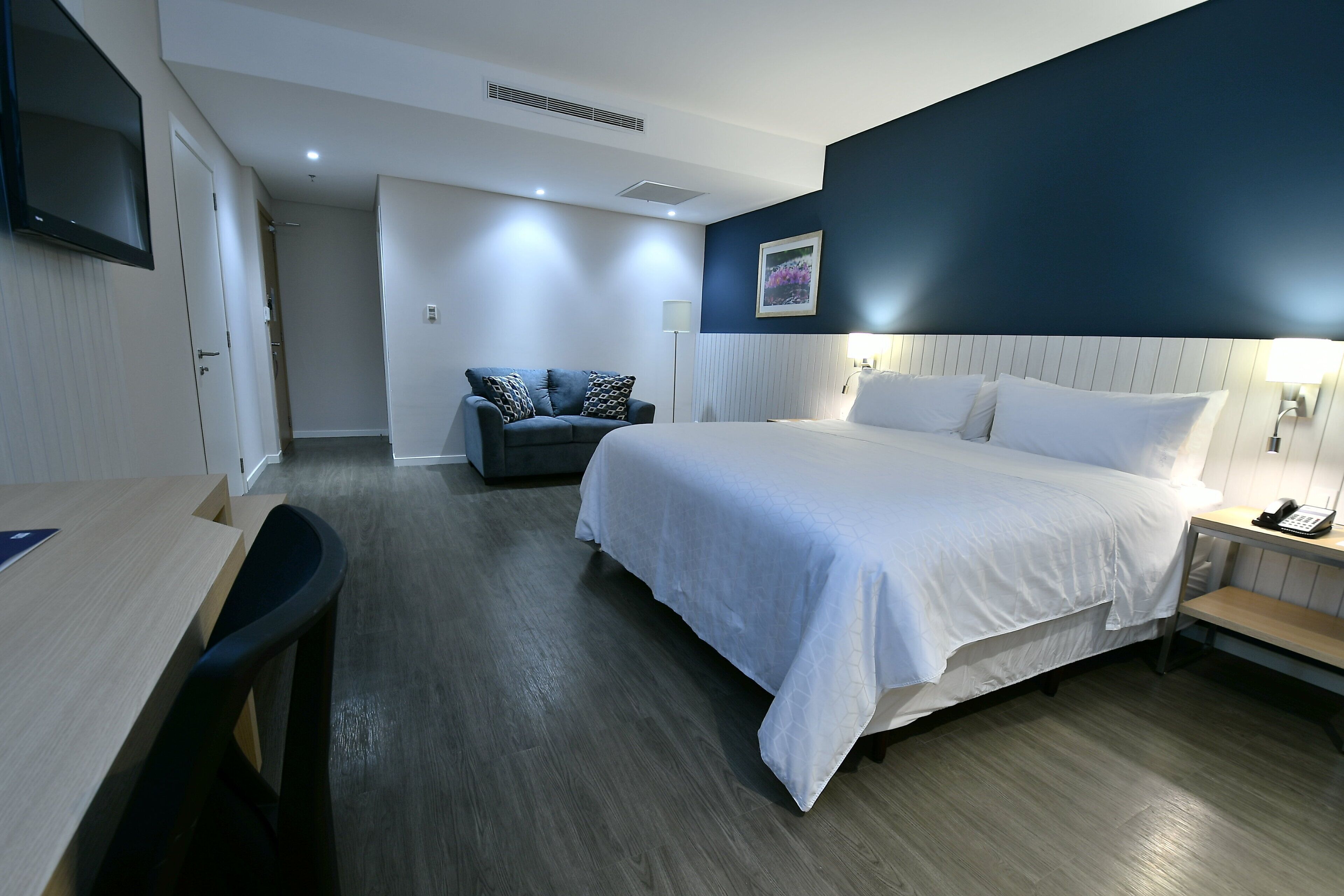 Ett hotellrum med en stor säng, ett skrivbord, en stol, en soffa och ett nattduksbord med en lampa.