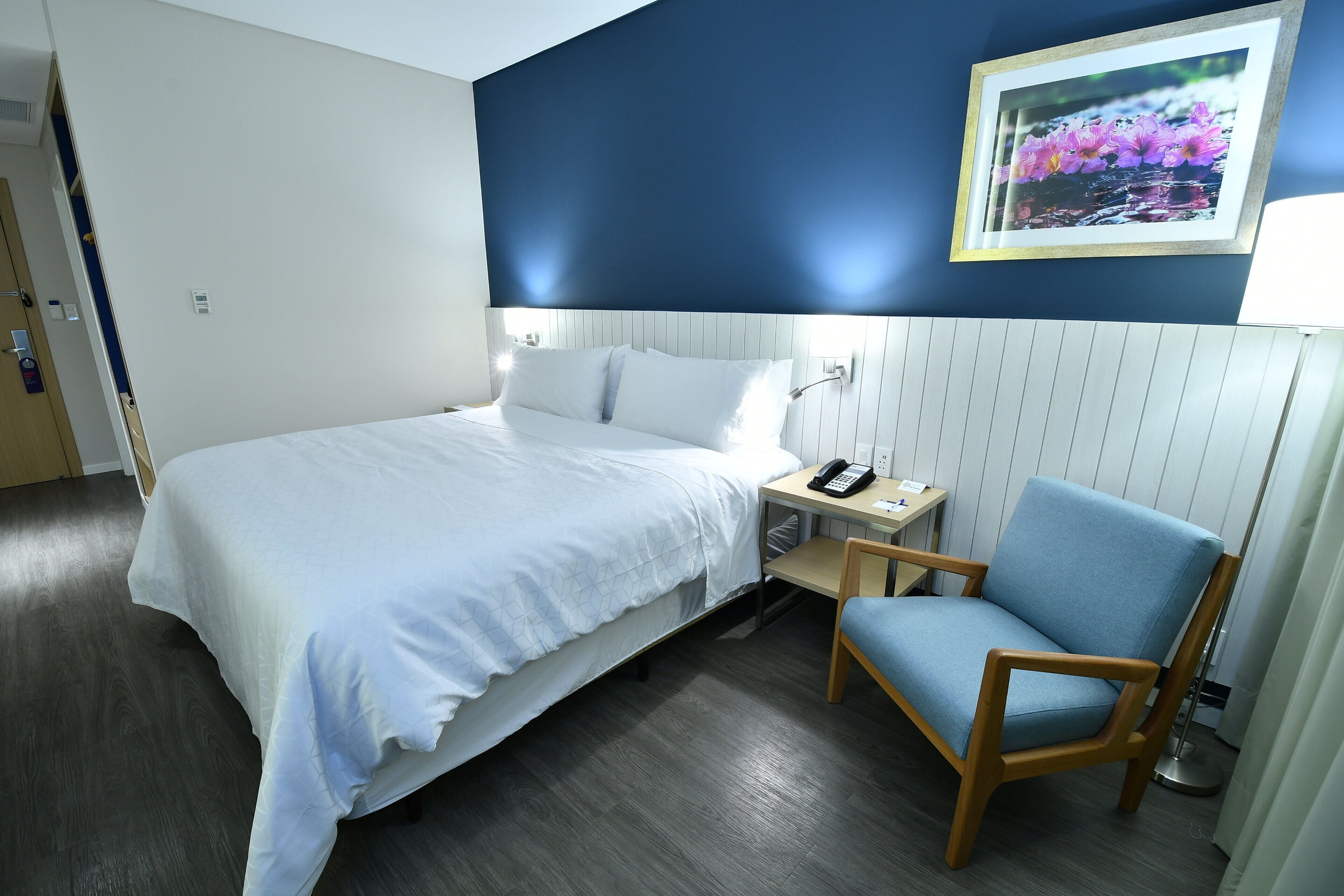 A hotel room with a bed, a chair, a desk, and a wall-mounted phone.