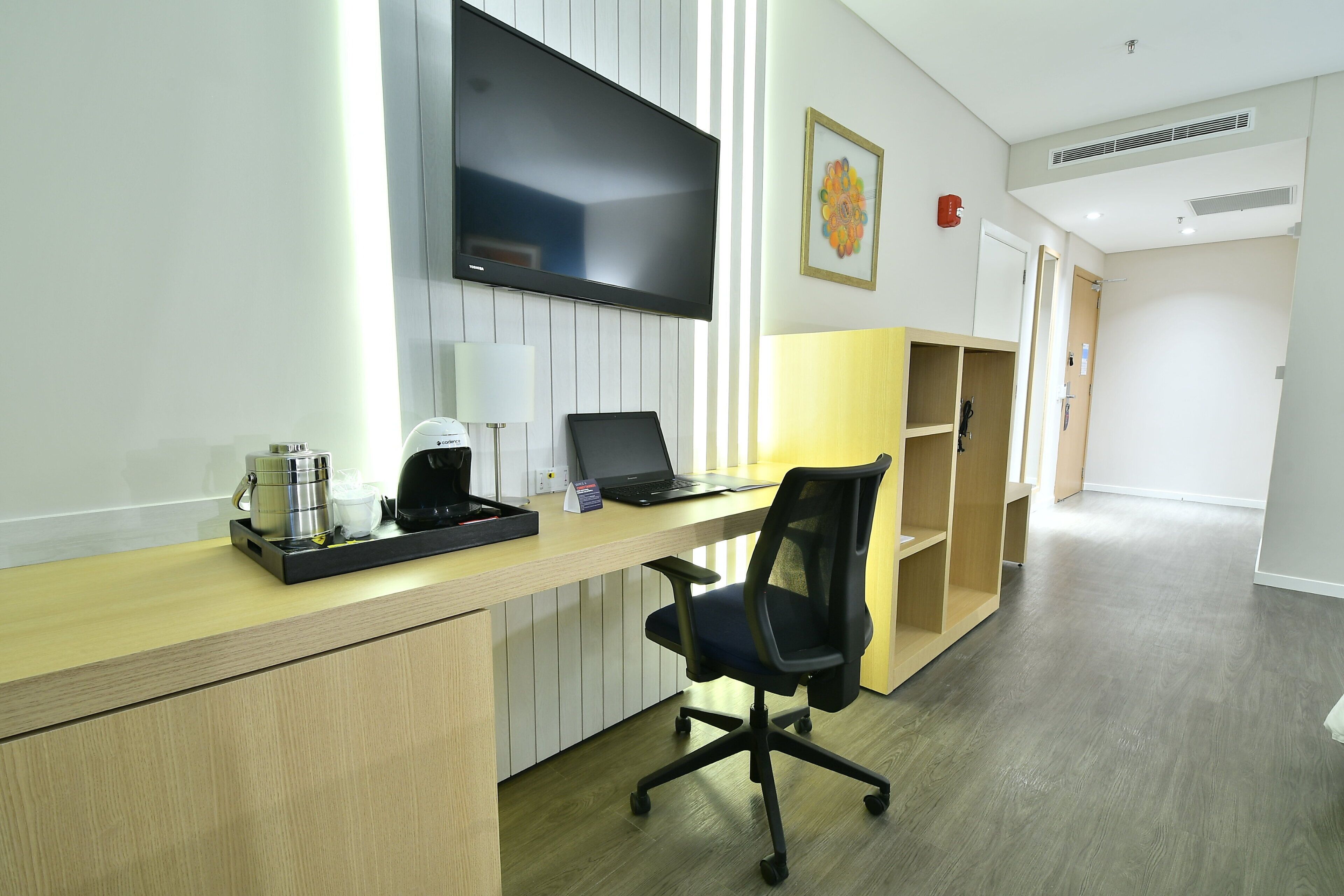A hotel room with a wooden desk, a black office chair, a flat-screen TV mounted on the wall, a coffee maker, and a laptop.