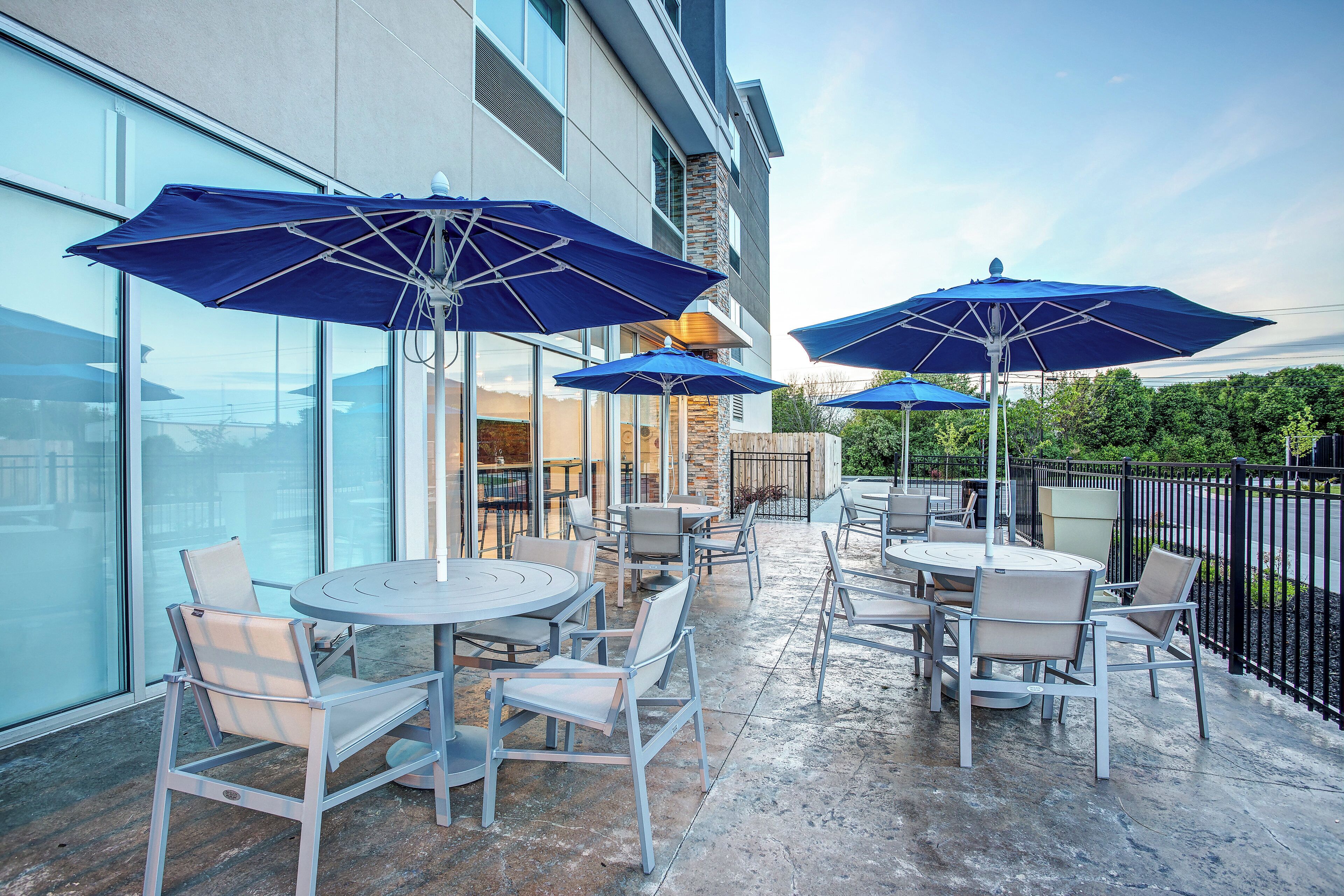 An outdoor seating area with tables and chairs, blue umbrellas, and a building with large windows.
