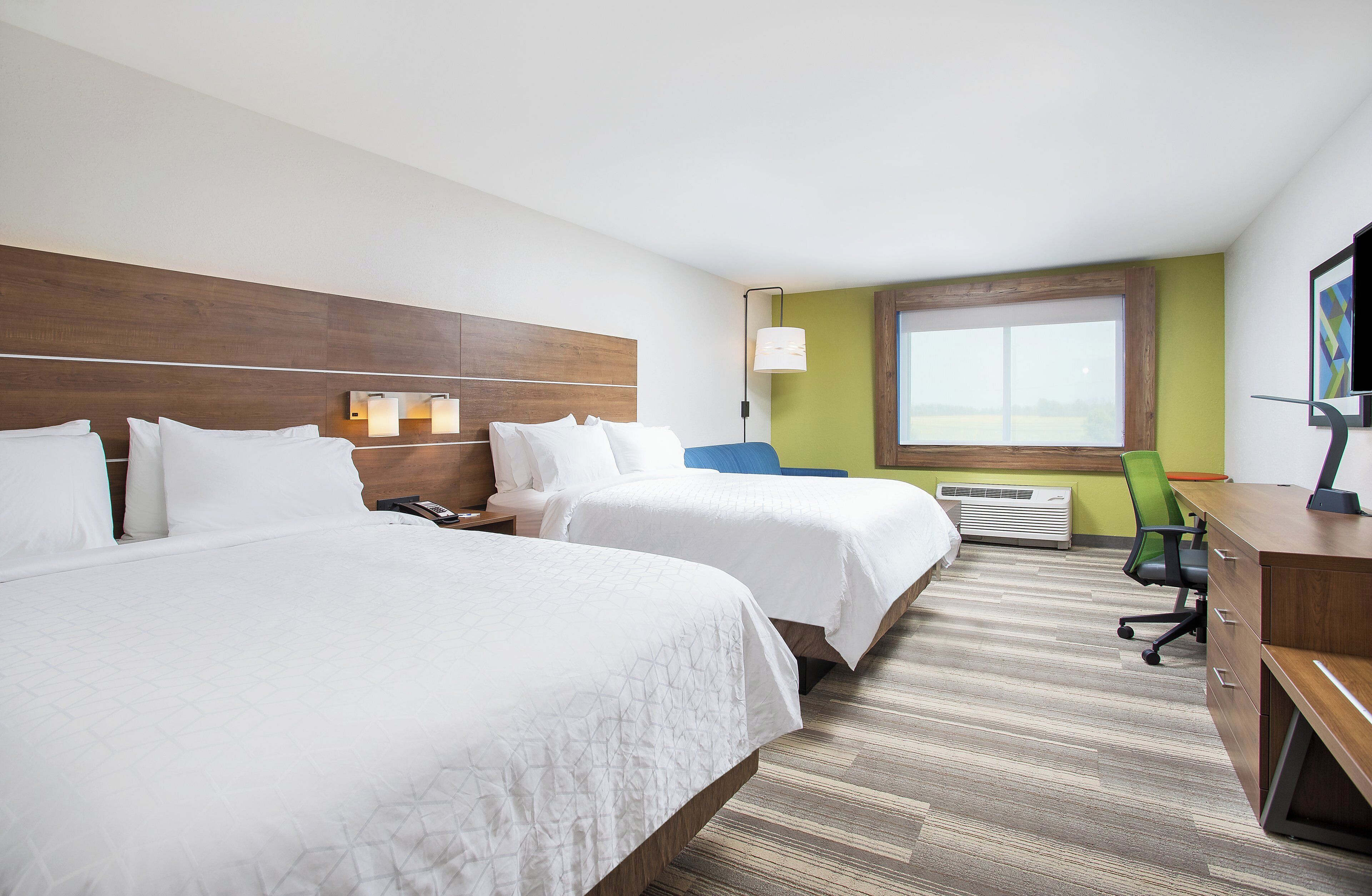 A hotel room with two beds, a desk, a chair, a window, and a wall-mounted TV.