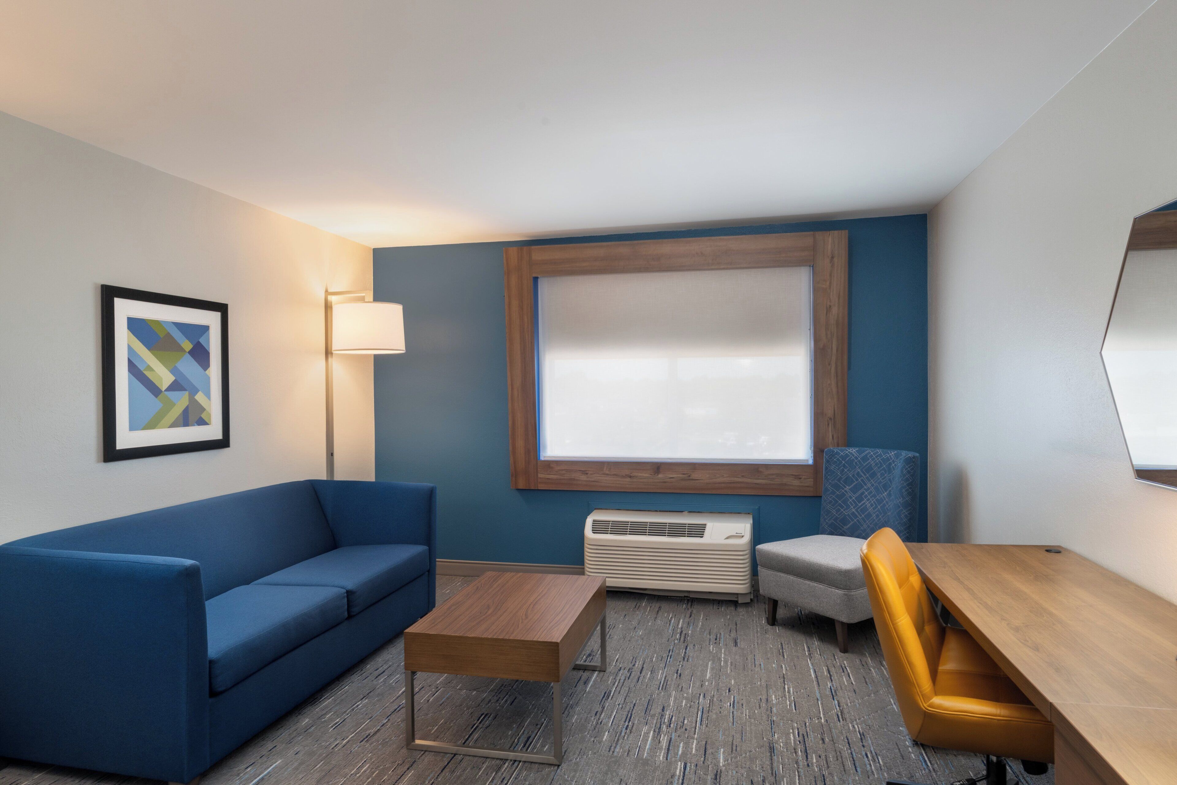 Una habitación de hotel moderna con un sofá azul, una mesa de centro de madera, un sillón pequeño y un escritorio con silla.