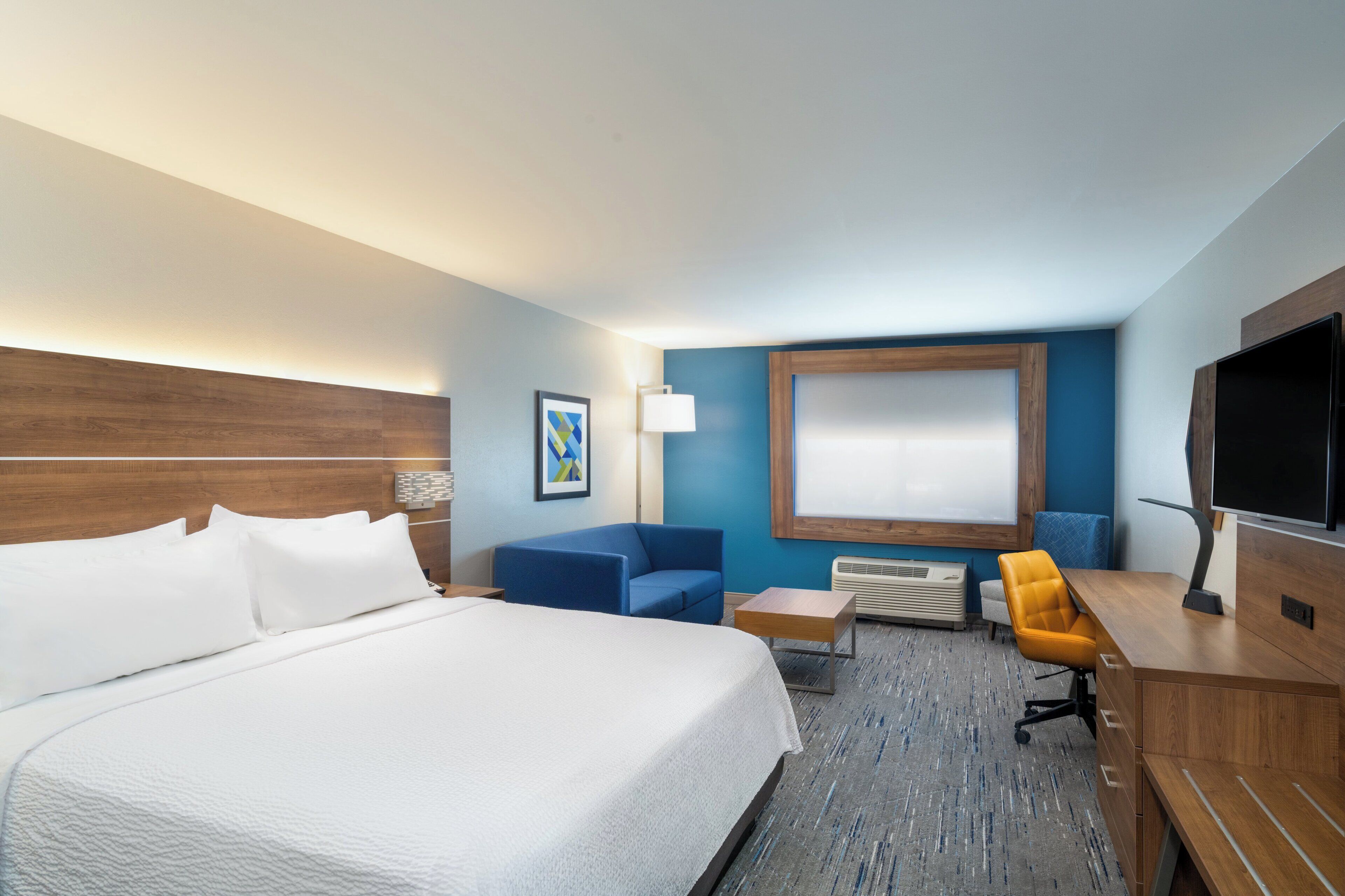 Ein Hotelzimmer mit einem großen Bett, einem Schreibtisch, einem Flachbildfernseher, einem blauen Sessel und einem kleinen Tisch.