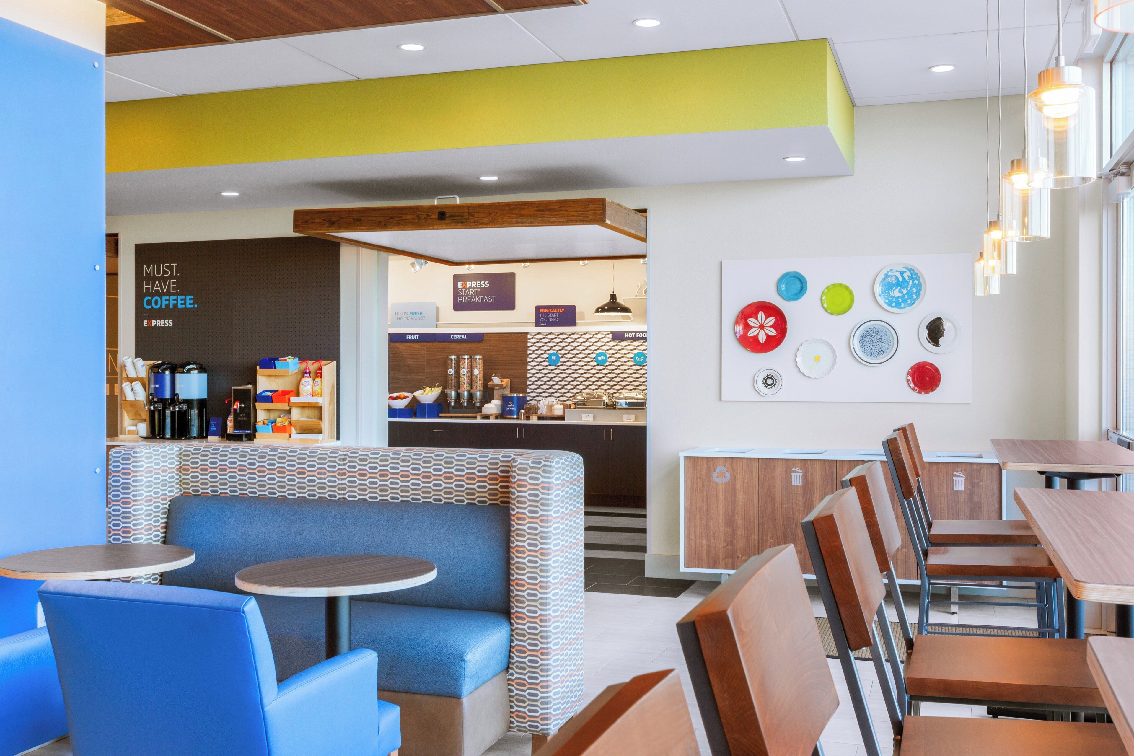 Un café moderne avec différents types de sièges, notamment des banquettes et des tables, un comptoir bien visible où est affiché le menu, et un mur décoré d’assiettes.