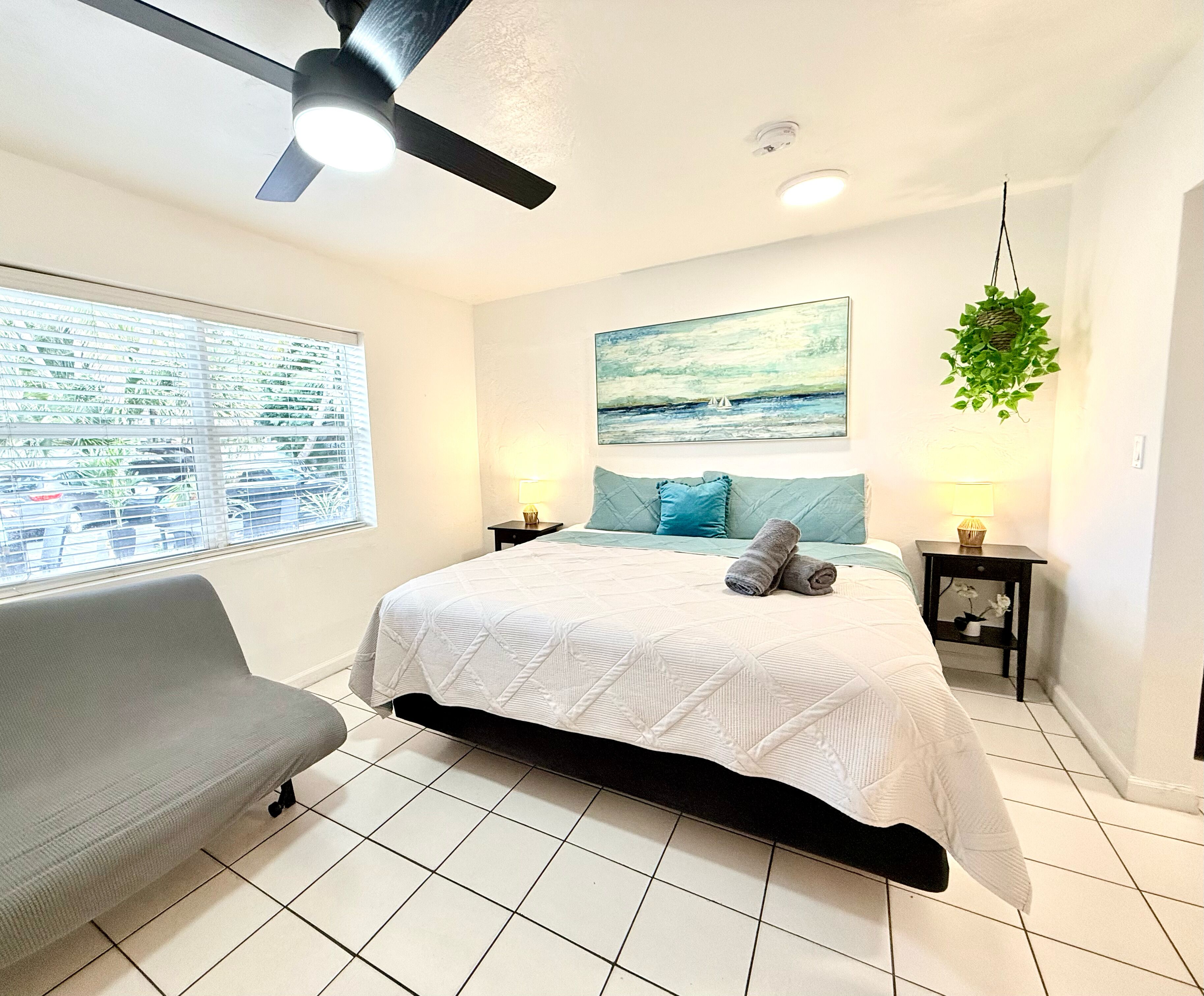 A bedroom with a bed, a ceiling fan, a window with blinds, a painting, a bedside table with a lamp, and a potted plant.
