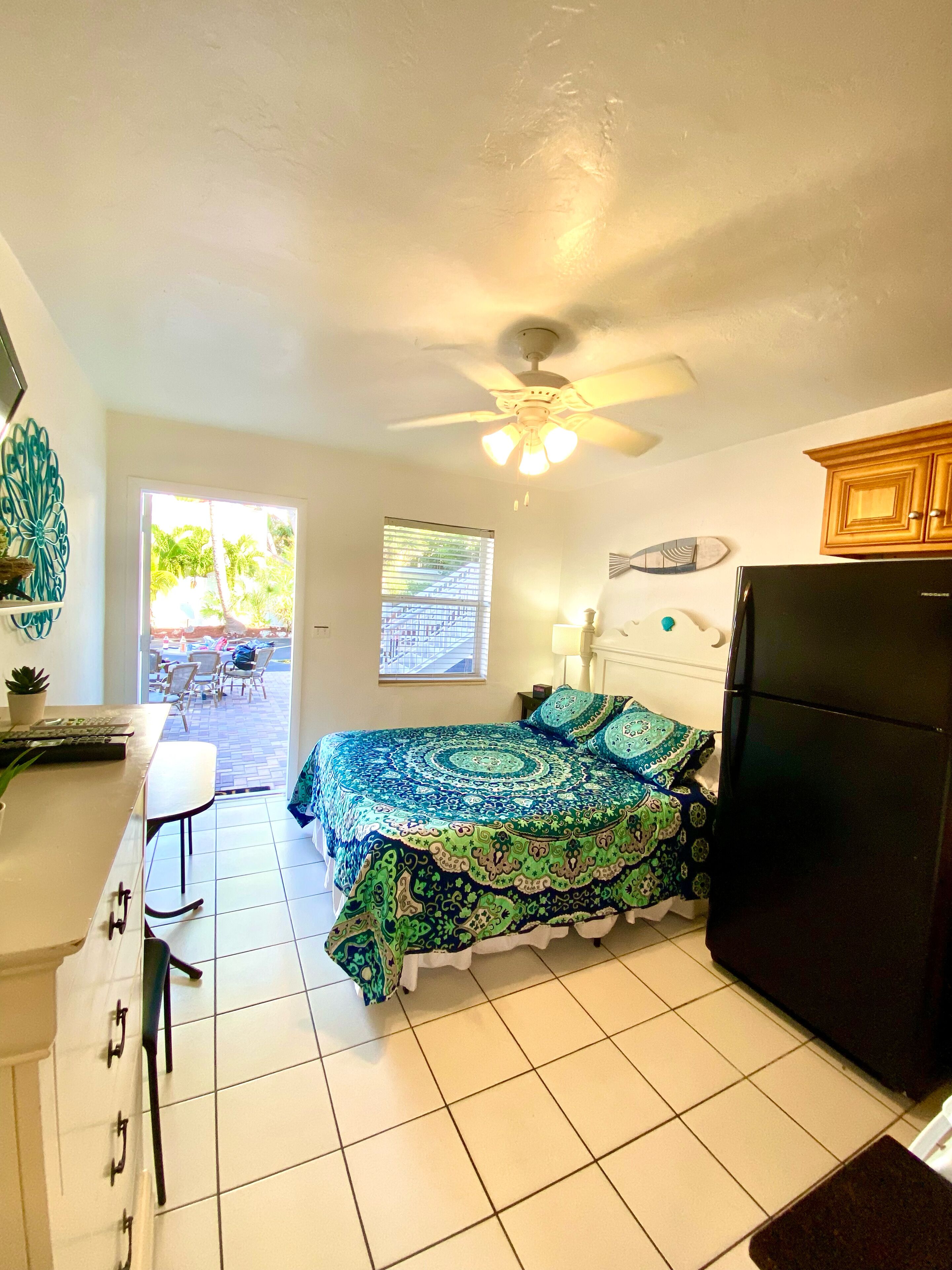 A compact bedroom with a bed, a ceiling fan, a refrigerator, and a small kitchenette.