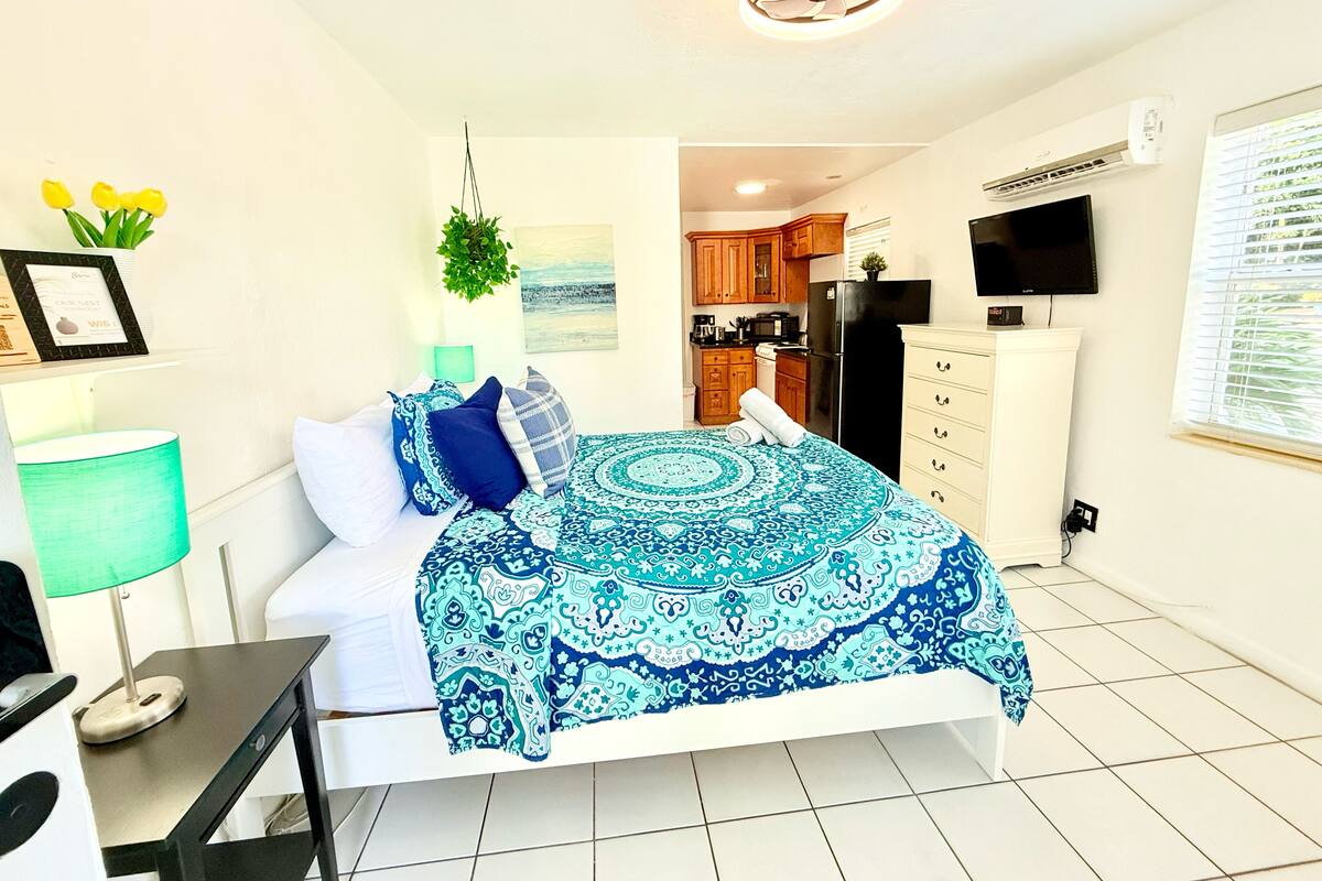 A bedroom with a bed, bedside table, lamp, and a view into the kitchen.