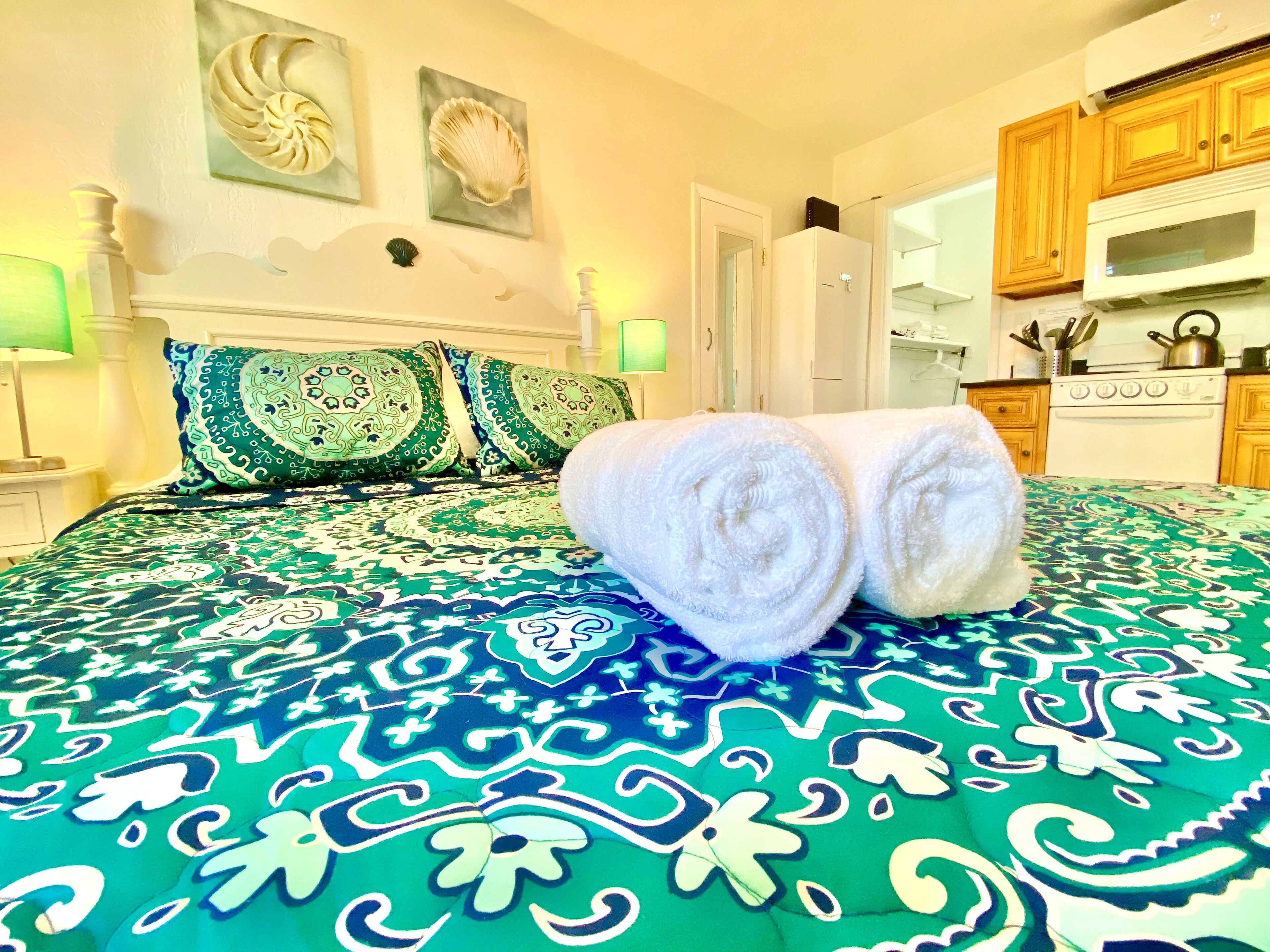 A hotel room with a bed, a green patterned bedspread, two decorative pillows, a white towel on the bed, a nightstand with a lamp, and a view of the kitchen.