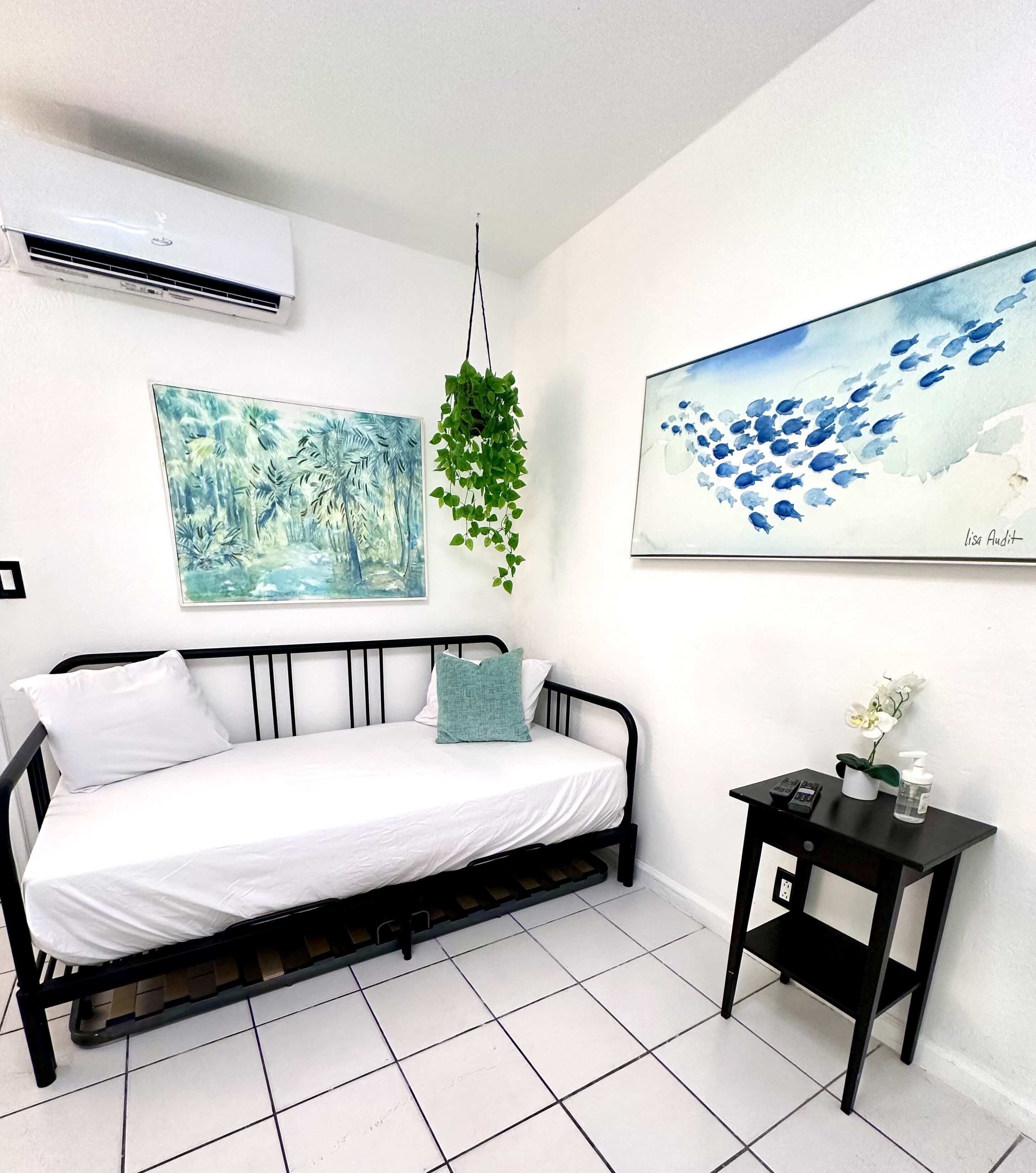 A bedroom with a bed, a small bedside table, a hanging plant, and two wall art pieces.