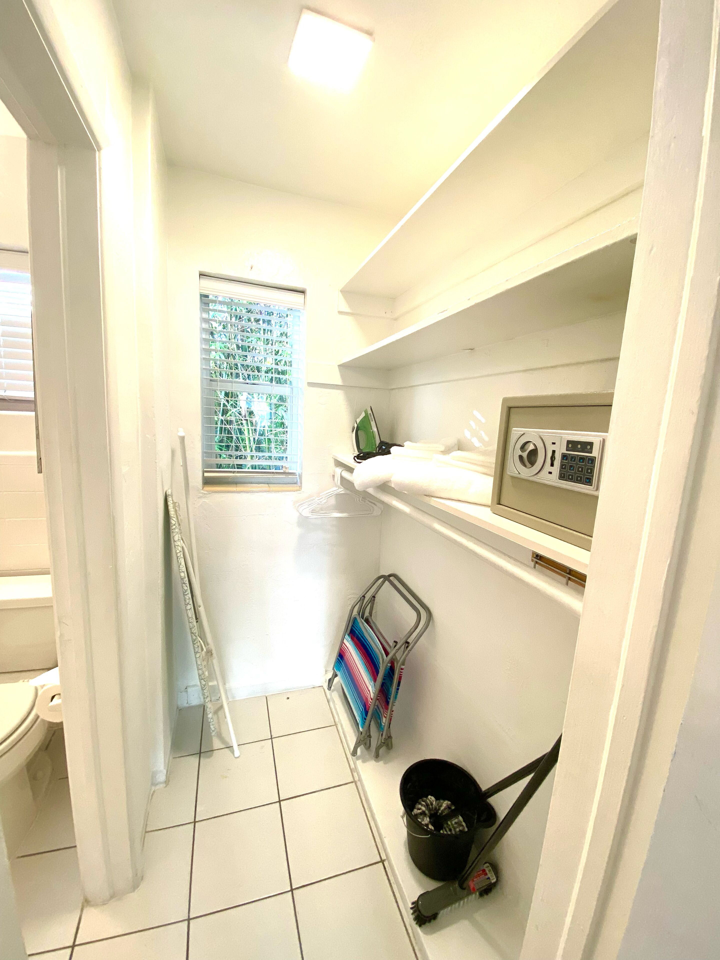 Ein kleines, weißes Badezimmer mit Toilette, Dusche, einem Fenster mit Jalousien und einem Regal mit Radio und Reinigungsmitteln.